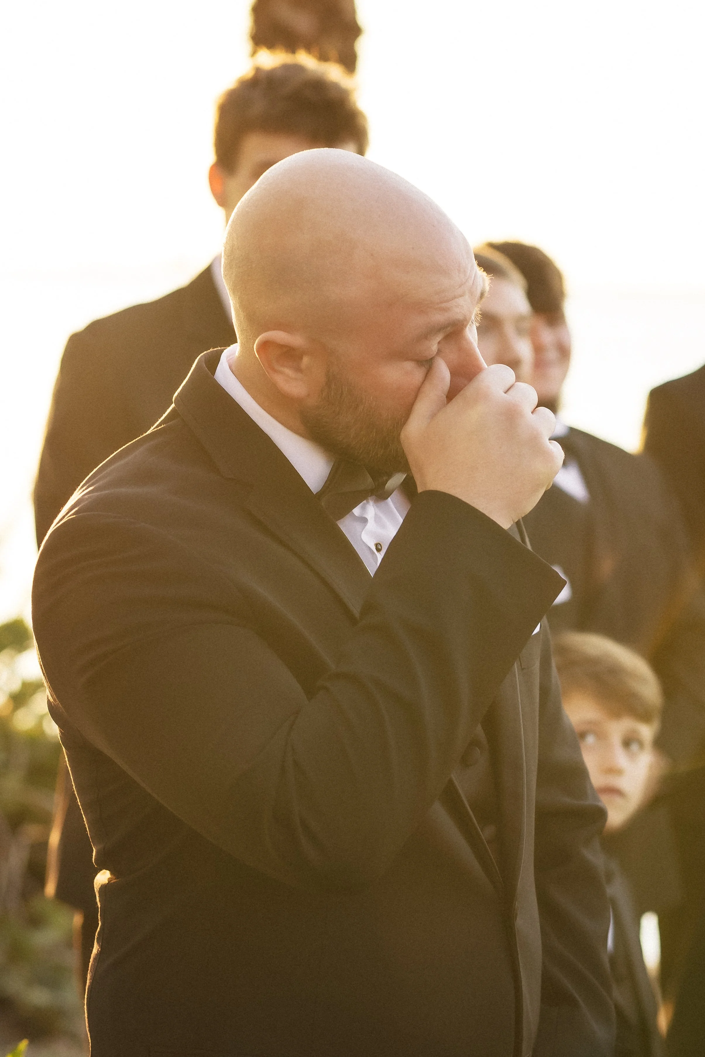 A man in a tuxedo with a beard and bald head is crying during a wedding ceremony outdoors, with a group of people dressed in formal attire behind him.