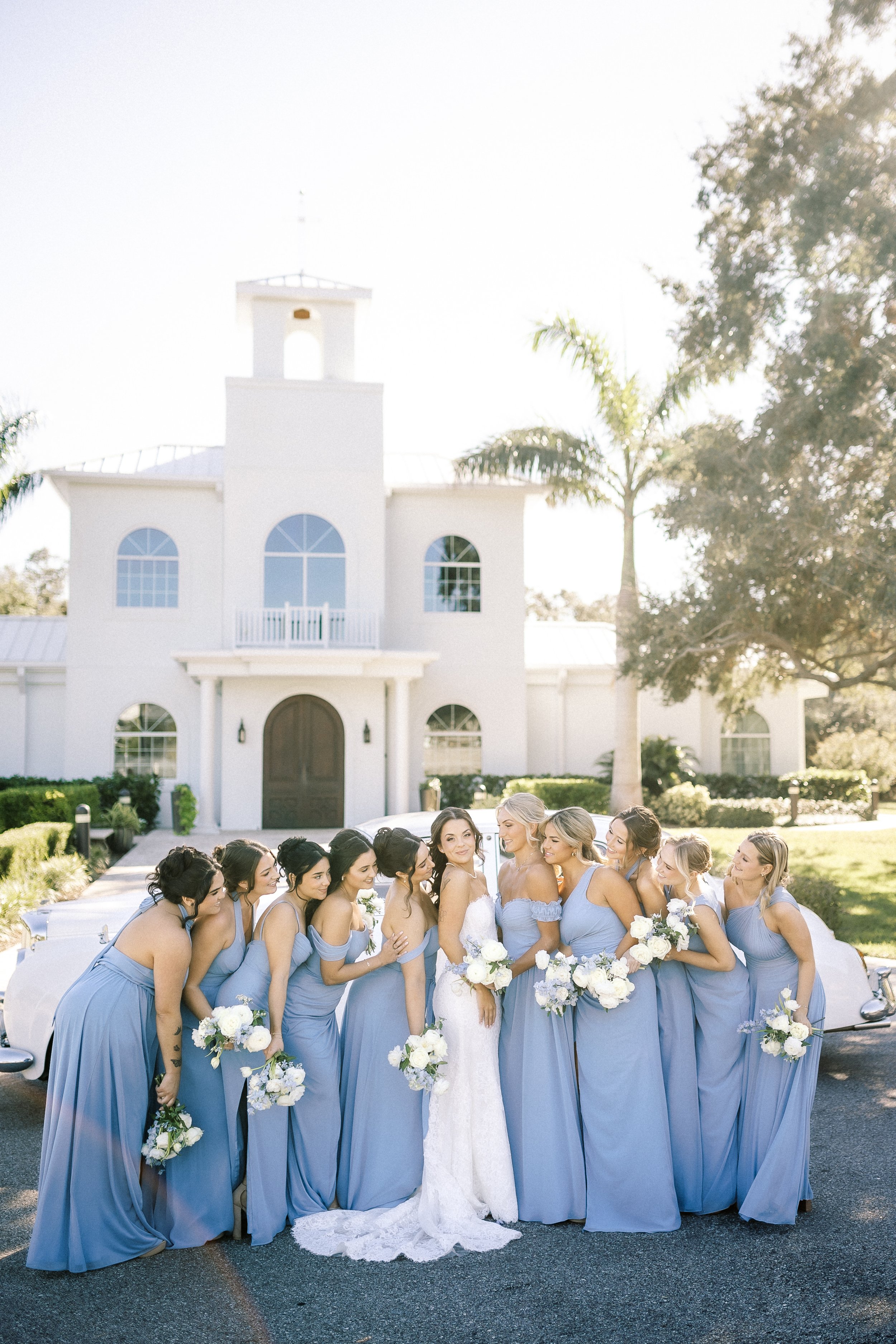 Bridal party standing outdoors in front of a white building, with the bride in white and bridesmaids in matching blue dresses holding bouquets.