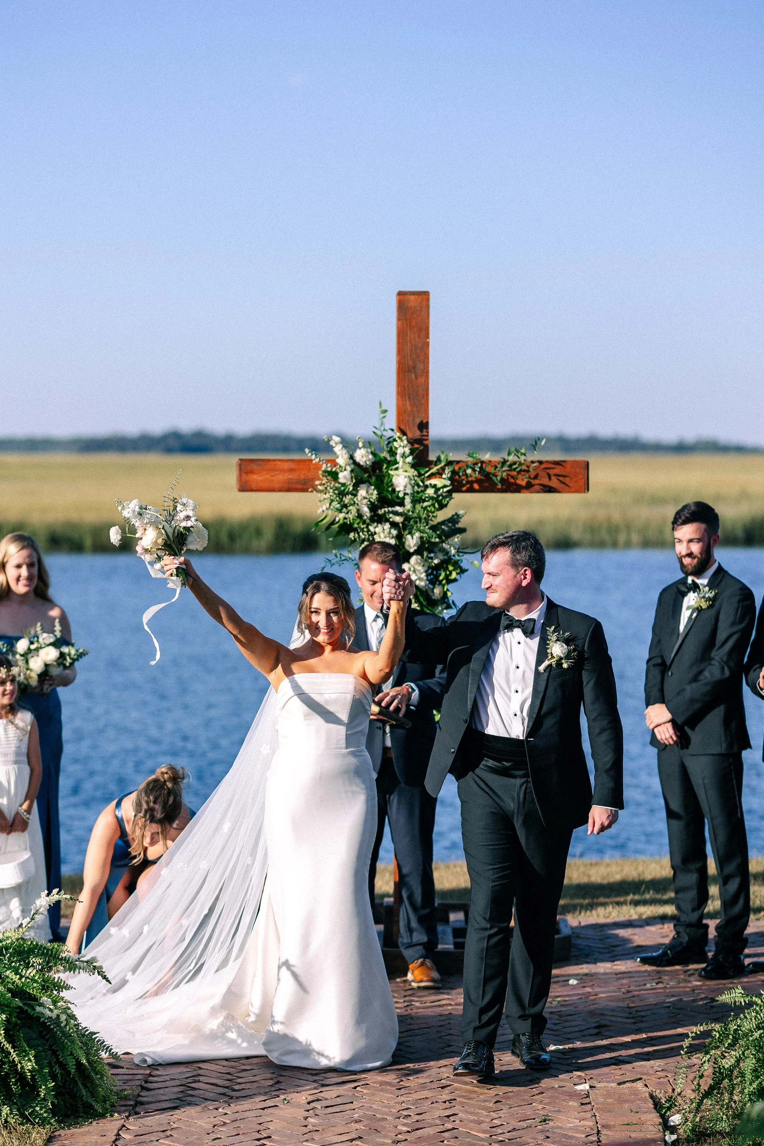 Bride and groom celebrating their wedding outdoors near a lake with a large wooden cross decorated with flowers in the background. The bride is holding a bouquet with outstretched arms, and the groom is holding her hand. Several guests are standing a