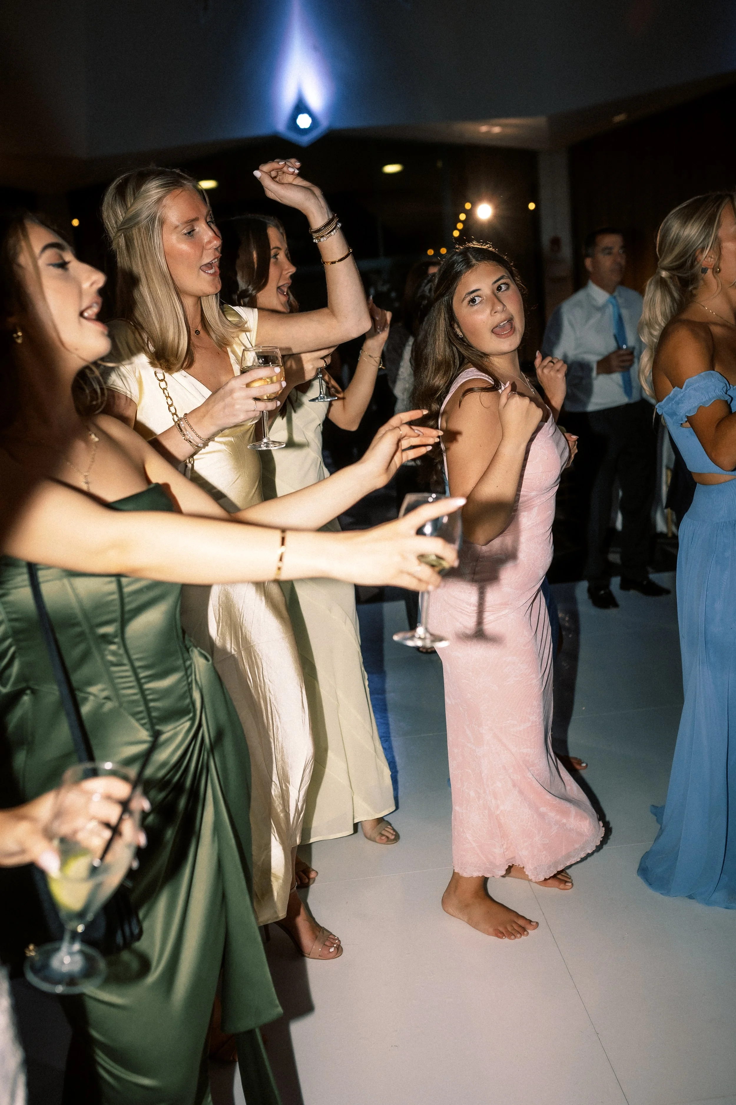 Group of women dancing and having fun at a party, some holding drinks, in a dimly lit venue.