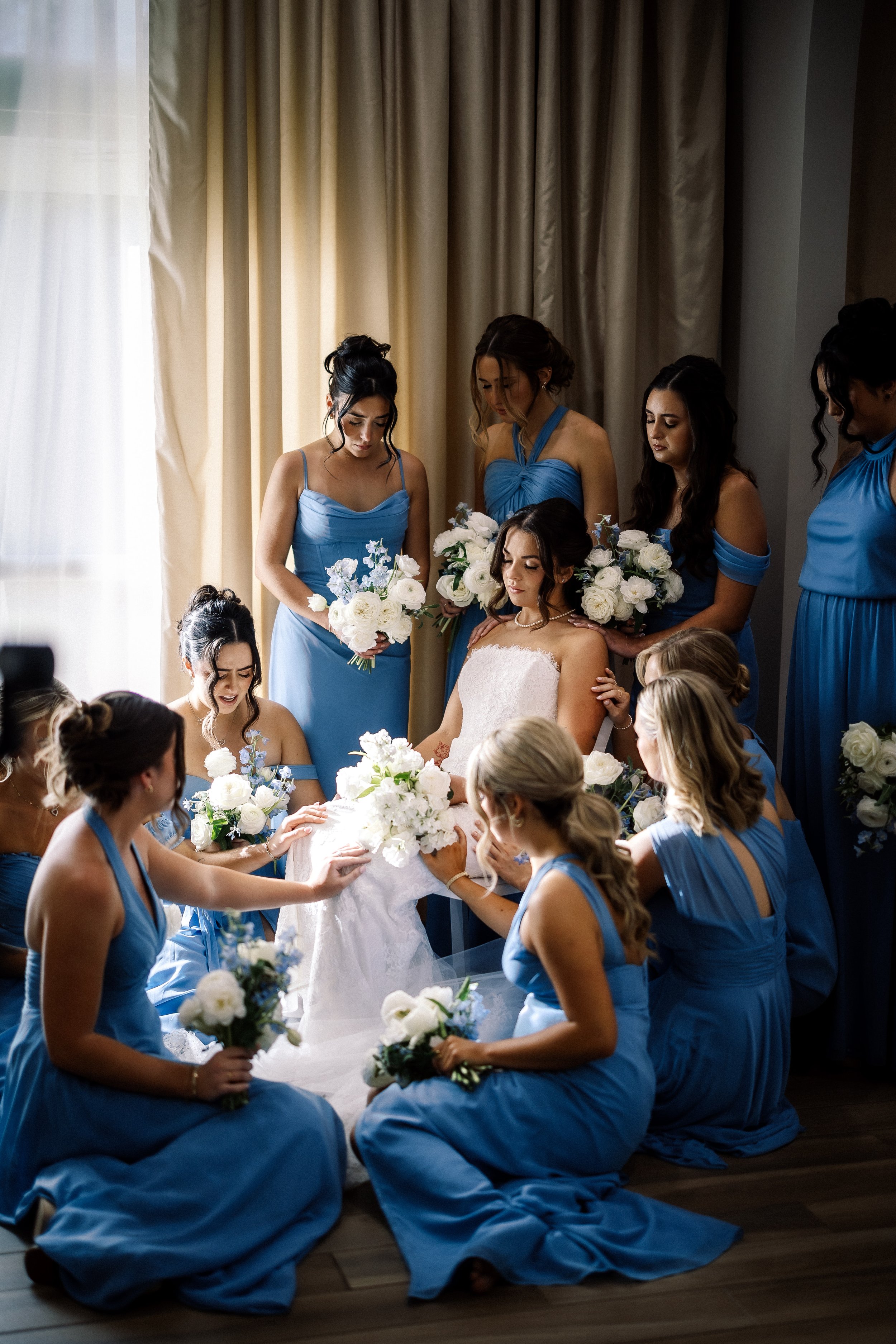 A bride surrounded by her bridesmaids and maid of honor in a wedding room, holding bouquets of white flowers, with a window and curtains in the background.