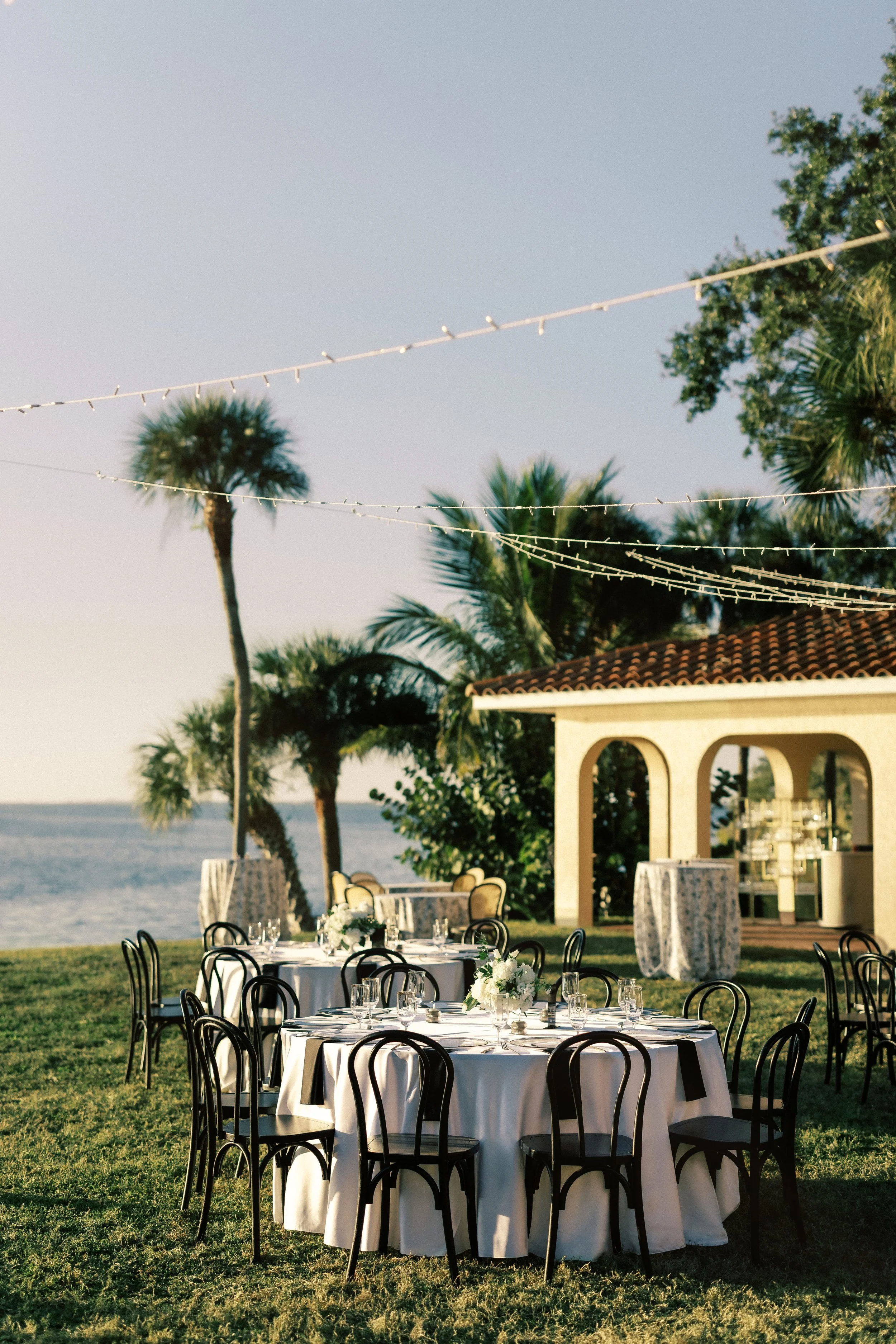 Outdoor dining setup with round tables, black chairs, floral centerpieces, on a grassy area near the ocean, with palm trees, a gazebo, and string lights.