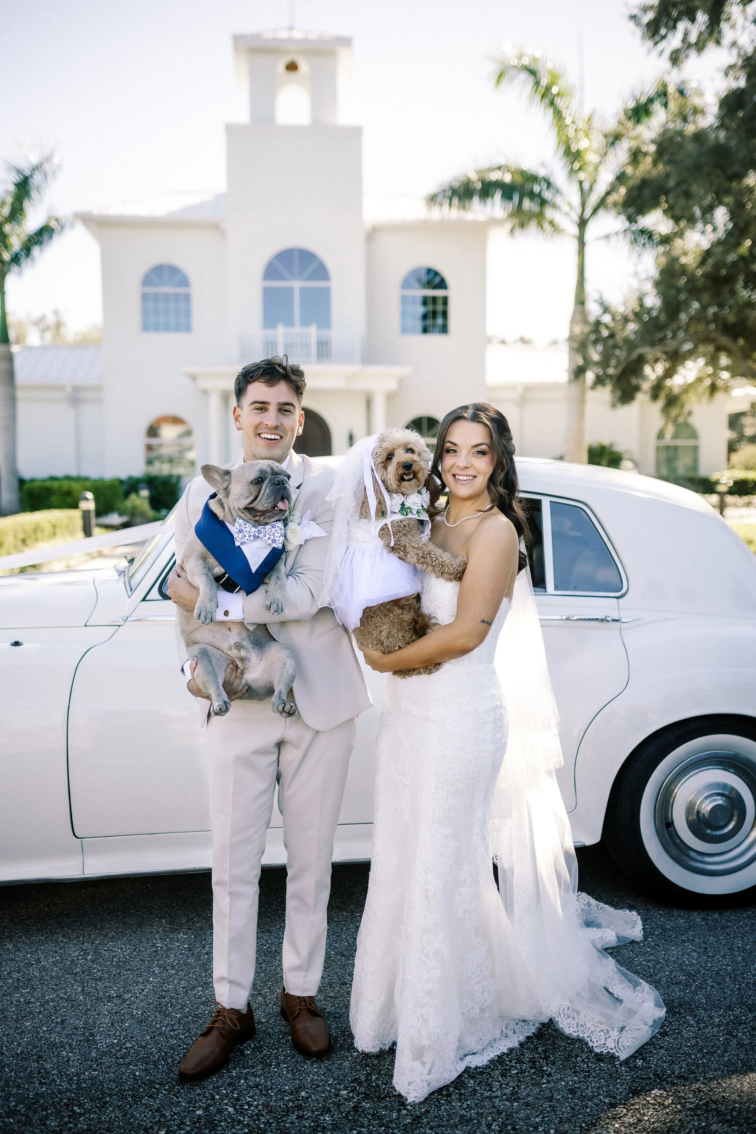 A newlywed couple in wedding attire standing in front of a white vintage car, holding their dogs, with a large white house and palm trees in the background.