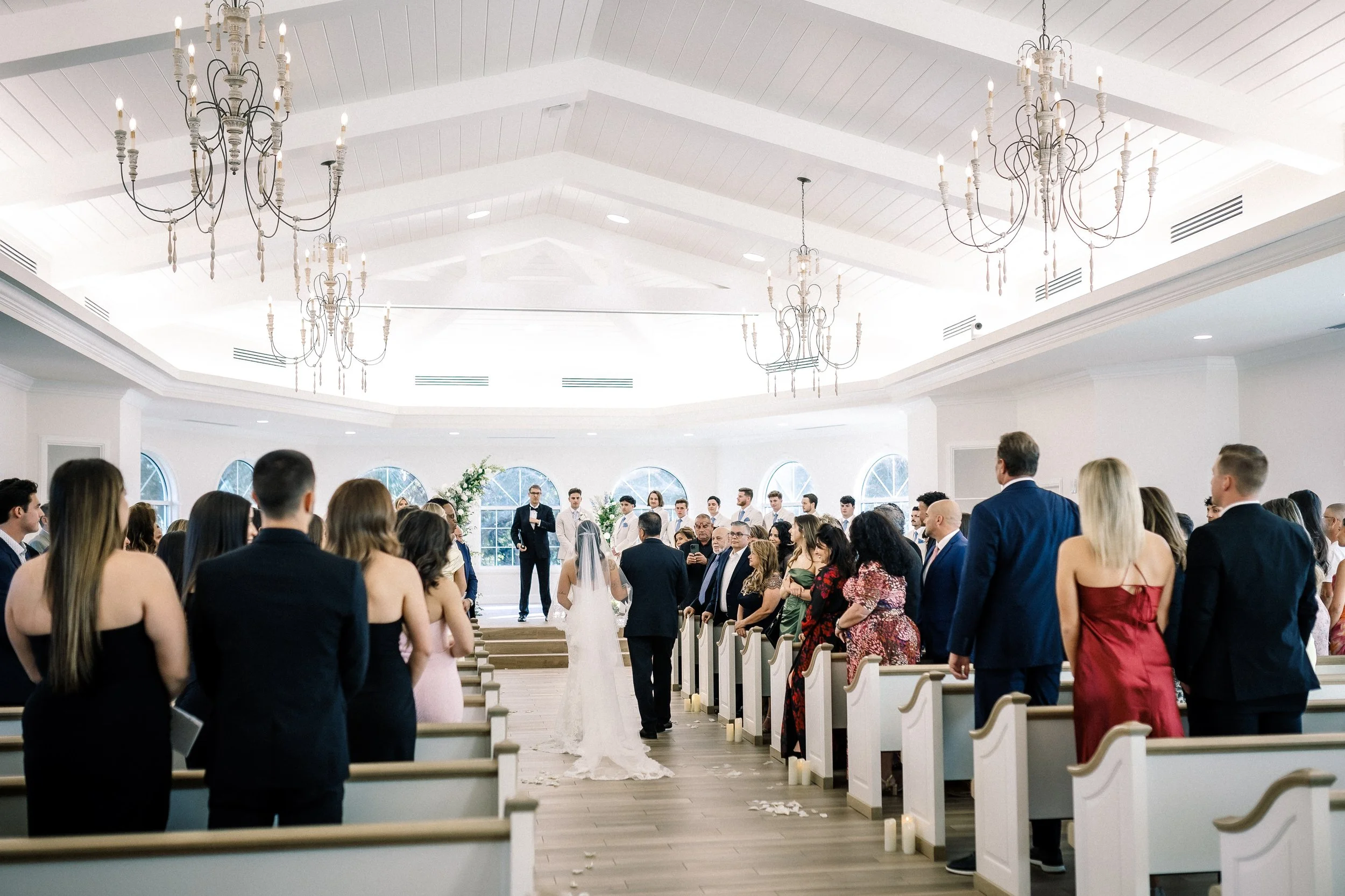 A wedding ceremony inside a bright, elegant chapel with chandeliers and large windows. The bride and groom walk down the aisle towards the altar as guests stand and watch.