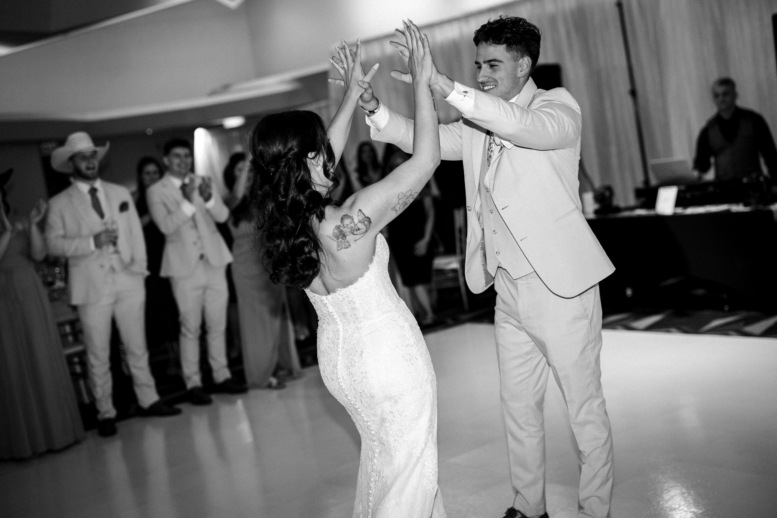 A bride and groom dancing at their wedding, surrounded by guests in formal attire, with the groom smiling and the bride raising her hands, black and white photo.