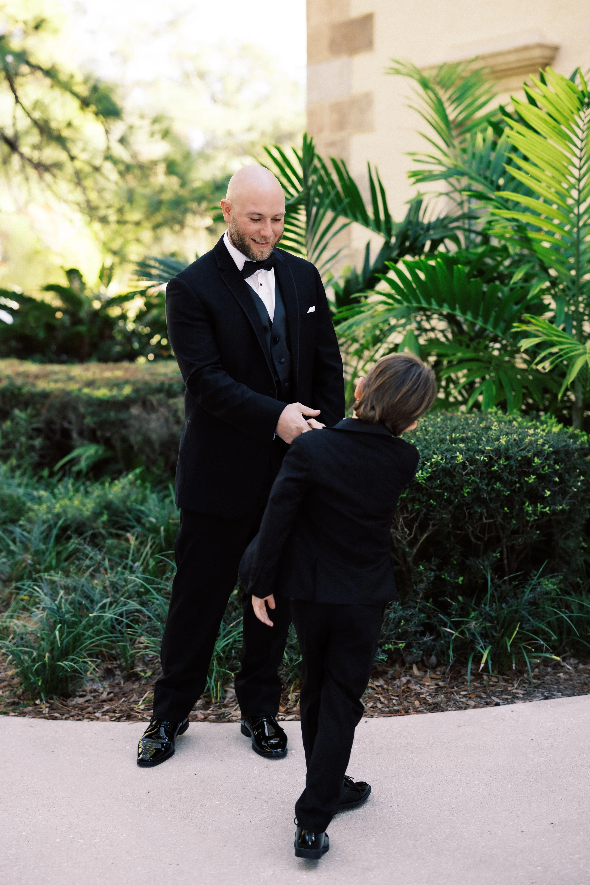 A man in a black tuxedo holding hands with a young boy in a black suit outdoors near green plants and a beige building.