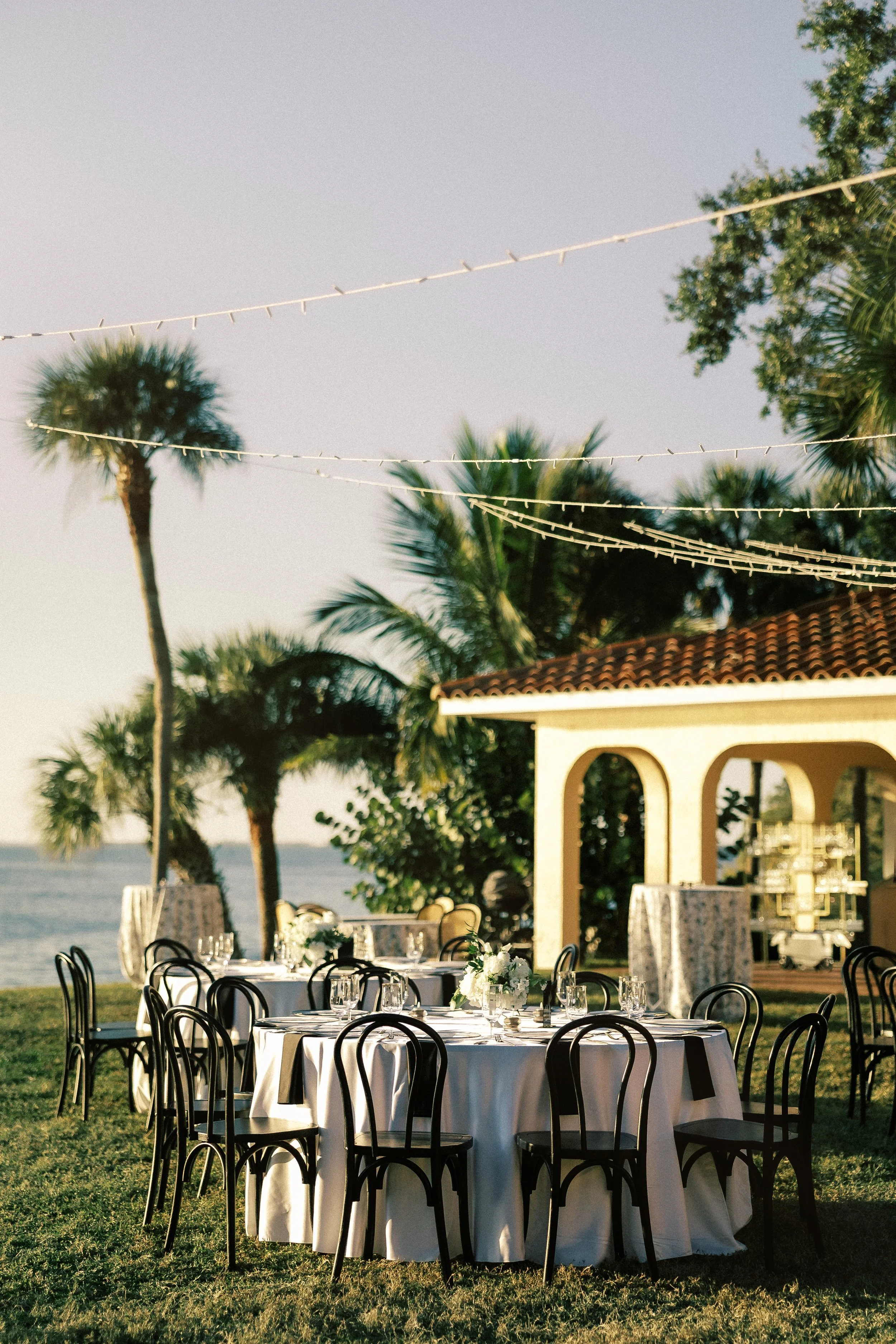 Outdoor event setup with round tables covered in white tablecloths, decorated with white floral centerpieces, surrounded by black chairs, near palm trees, with a building featuring arched openings and string lights overhead, overlooking a body of wat