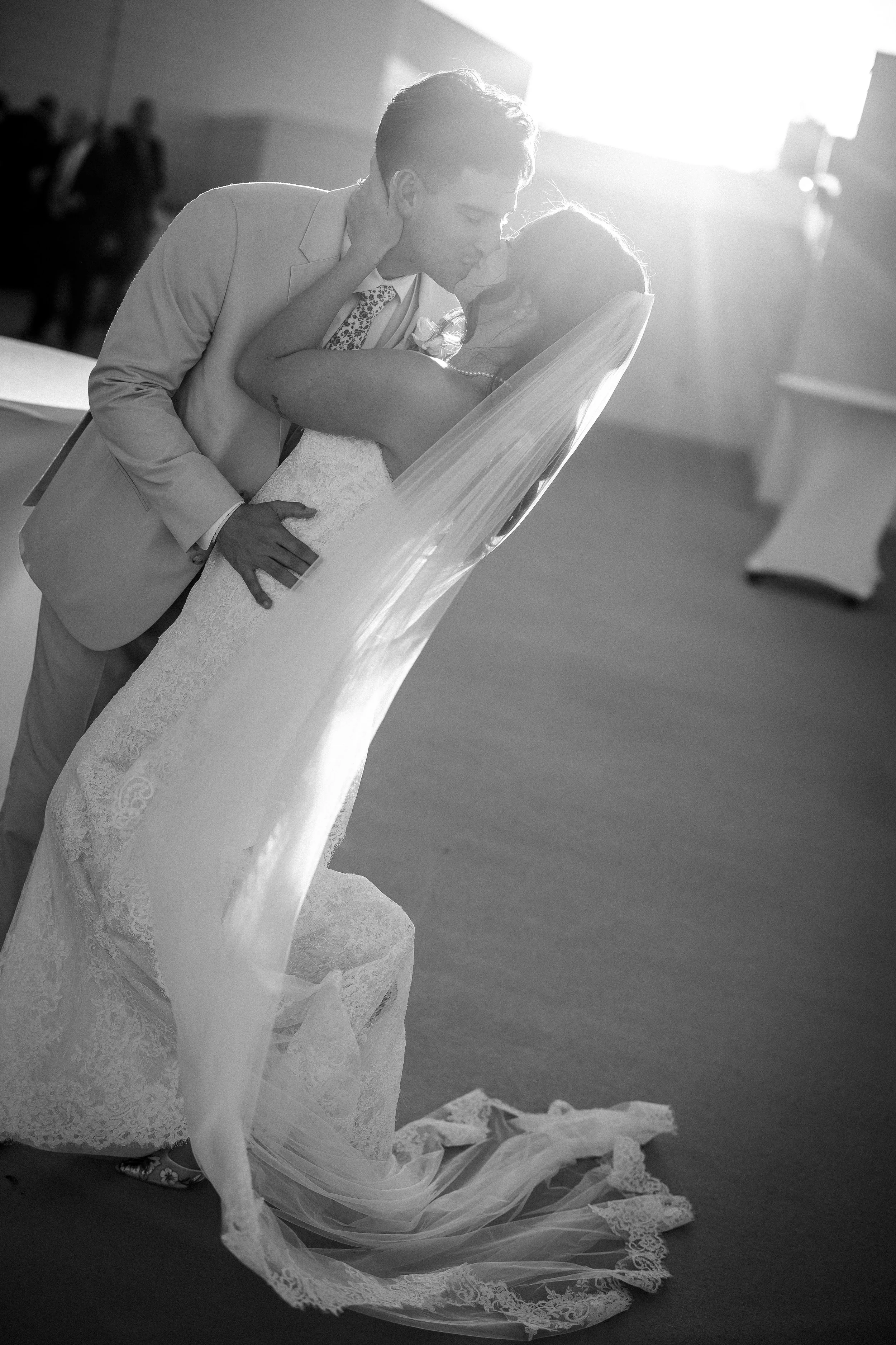 A newlywed couple sharing a kiss during their wedding ceremony, with the bride in a lace wedding gown and veil, and the groom in a suit, in a well-lit indoor setting.