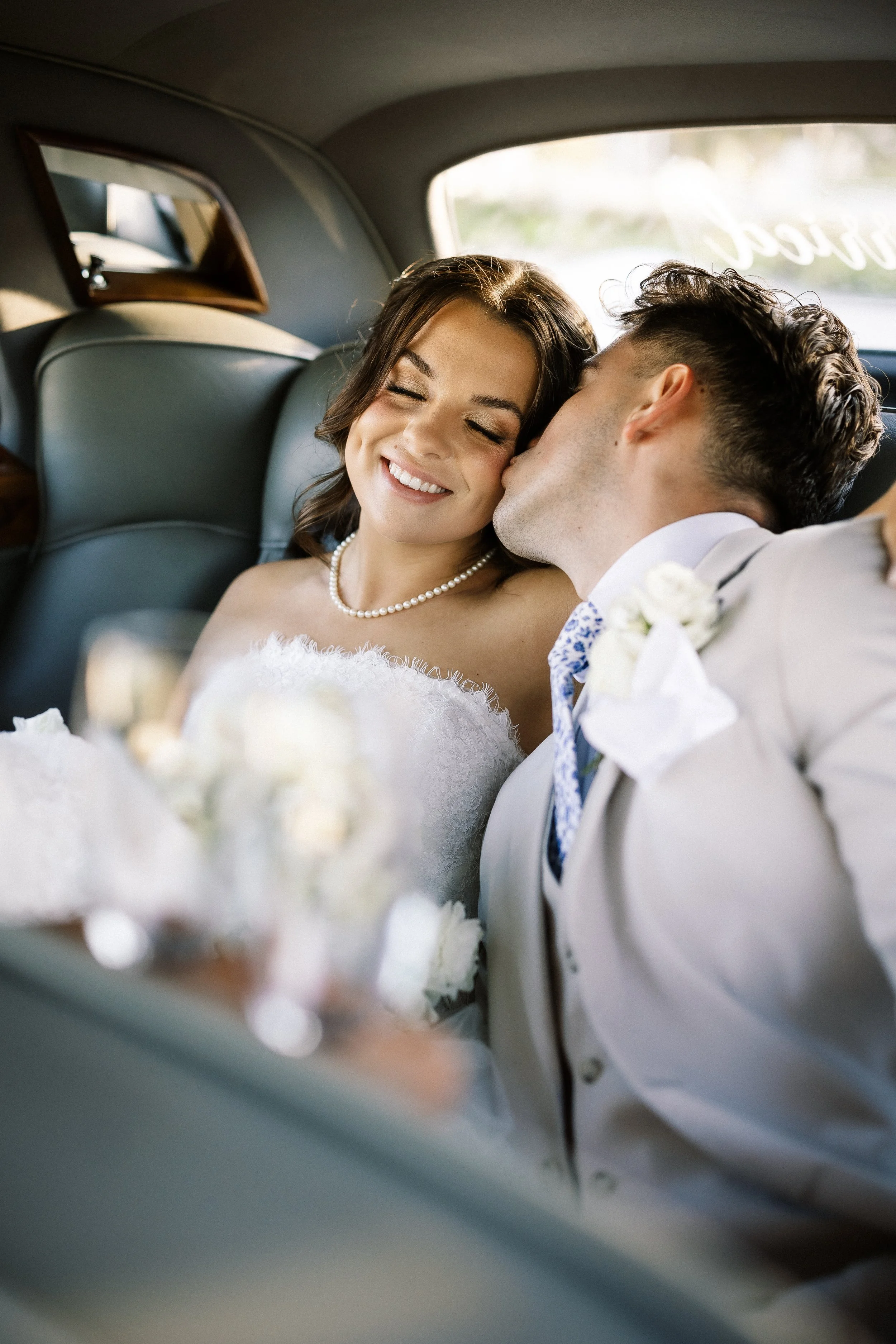 A bride and groom sitting together in a car, with the groom kissing the bride's temple. The bride is smiling with her eyes closed, wearing a white lace dress and pearl necklace. The groom is in a light suit with a boutonniere, and they appear happy a