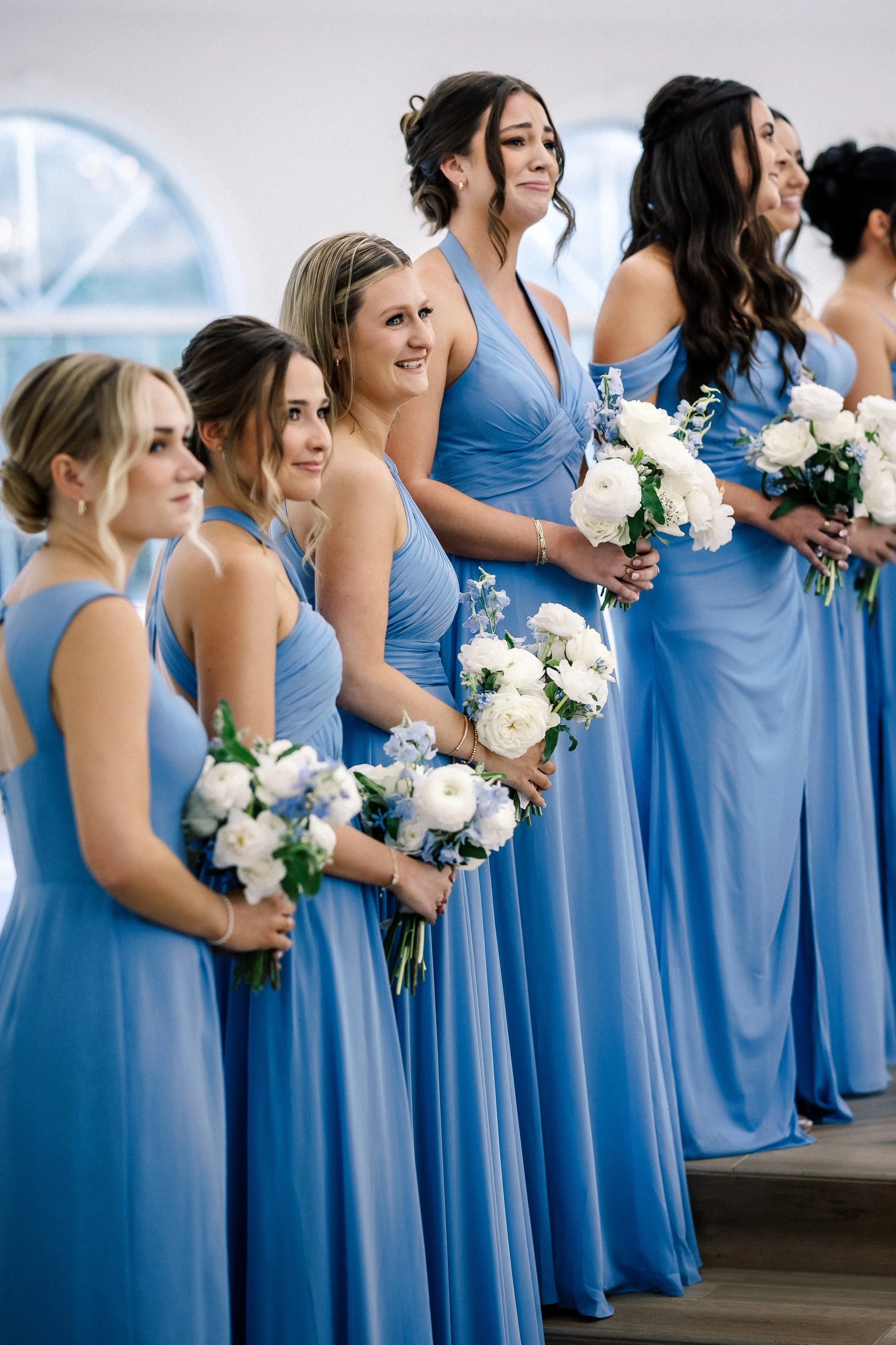 A group of bridesmaids standing in a row indoors, dressed in matching long blue dresses, holding white floral bouquets.