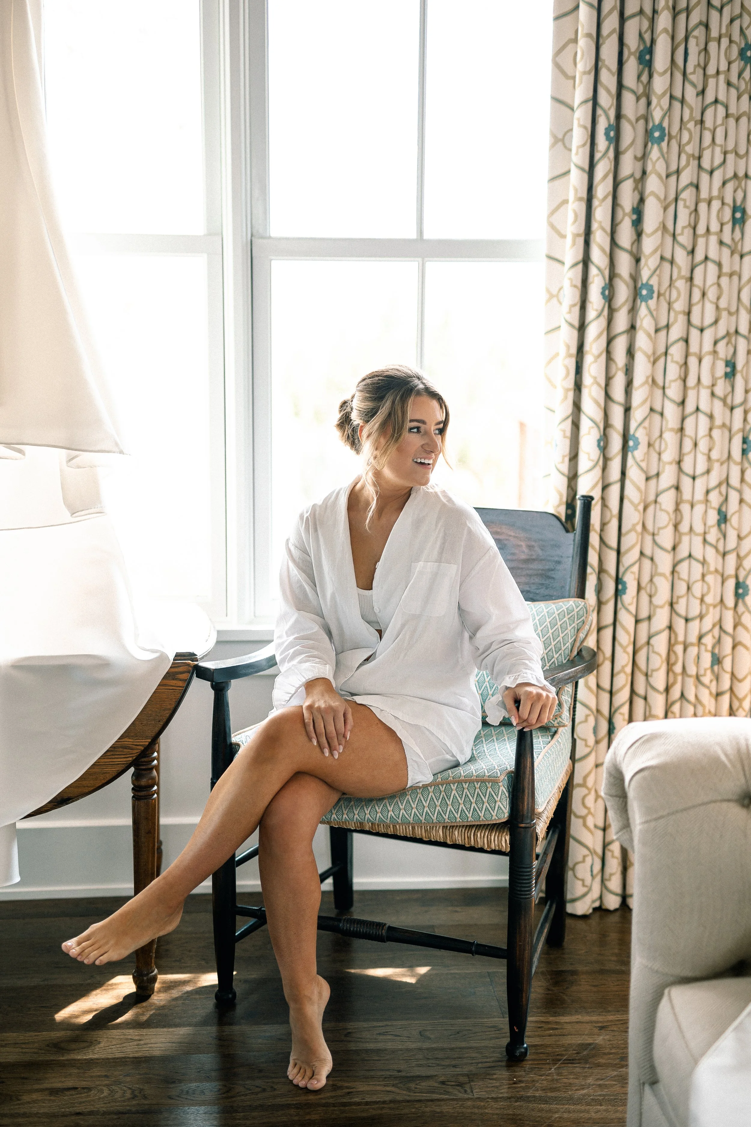 A woman dressed in a white robe sitting on a cushioned chair near a window, smiling and looking away.