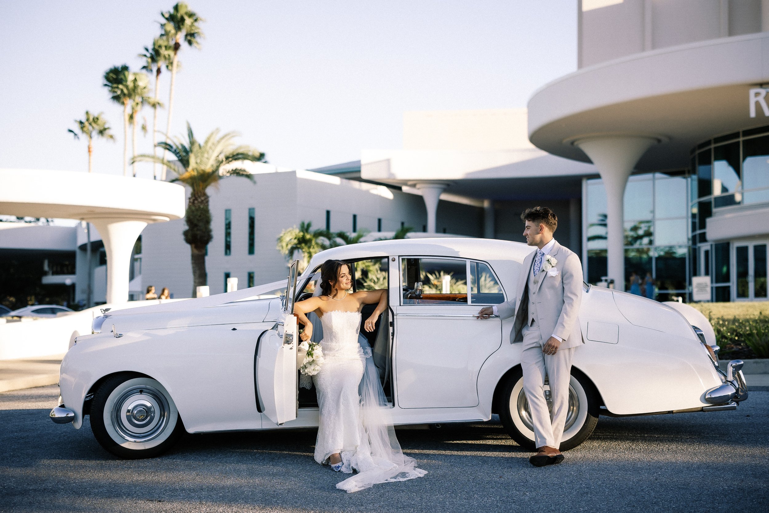 A bride and groom on their wedding day, standing outside next to a vintage white car. The bride is sitting inside the car holding a bouquet, and the groom is standing outside, dressed in a light beige suit, holding the door handle, smiling at her. Th