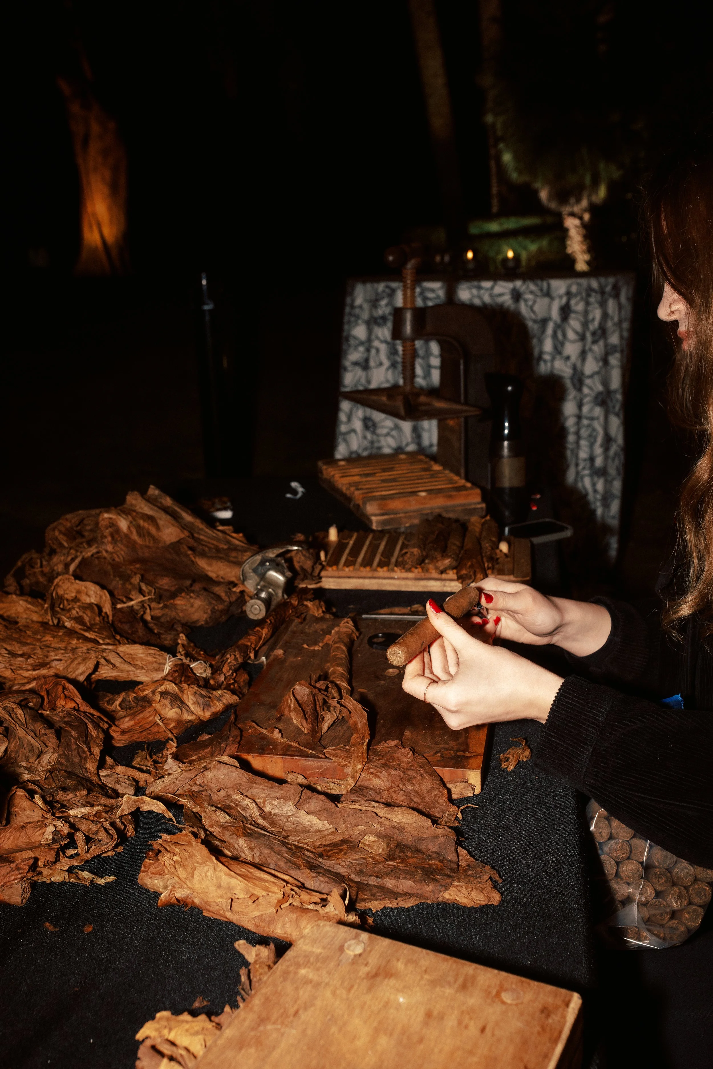 A person handling cigars at a cigar station, with cigars in various stages of preparation, on a black surface, in a dimly lit room.