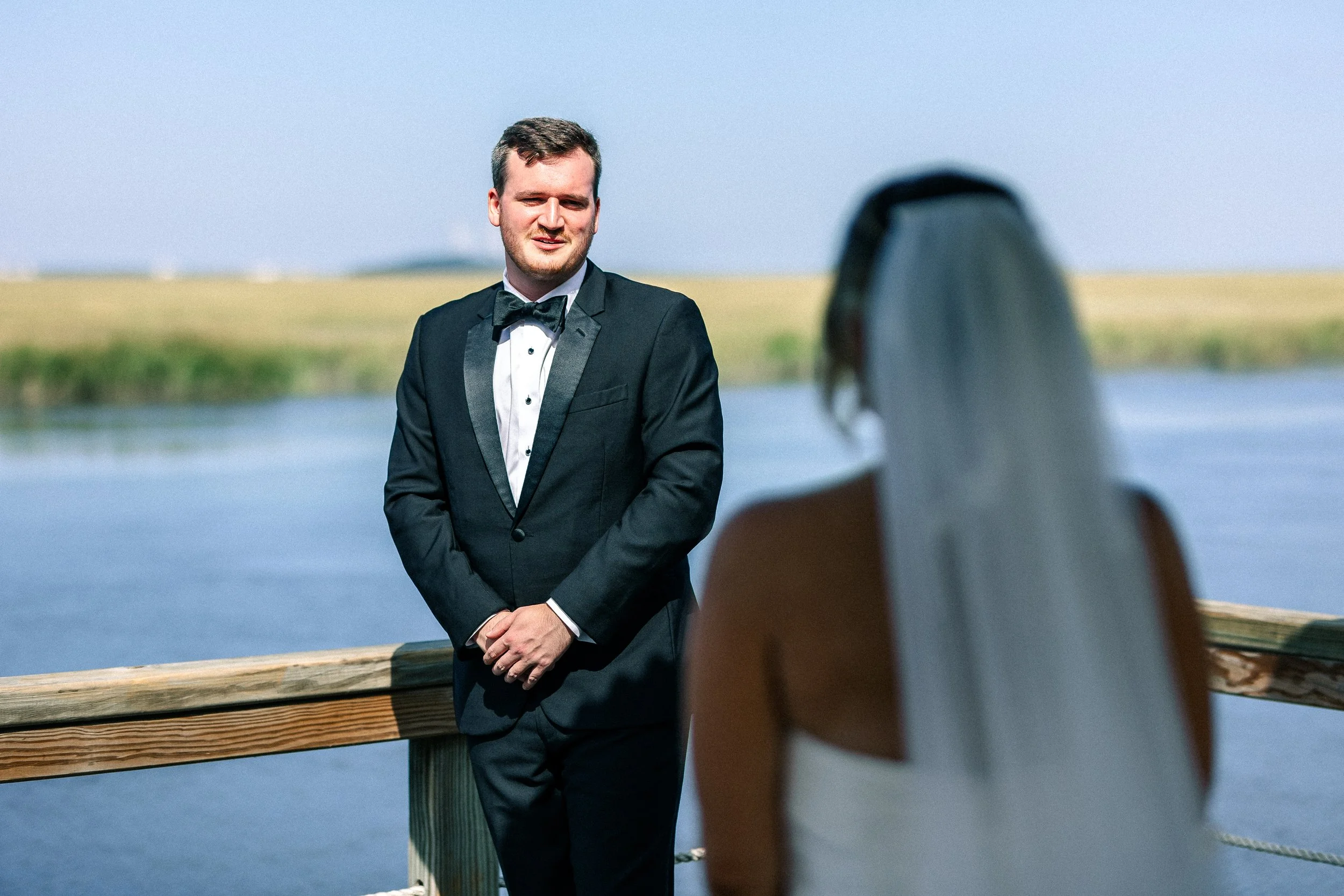 A man in a tuxedo standing outdoors in front of a body of water, with a woman sitting in foreground with long hair, on a wooden deck.