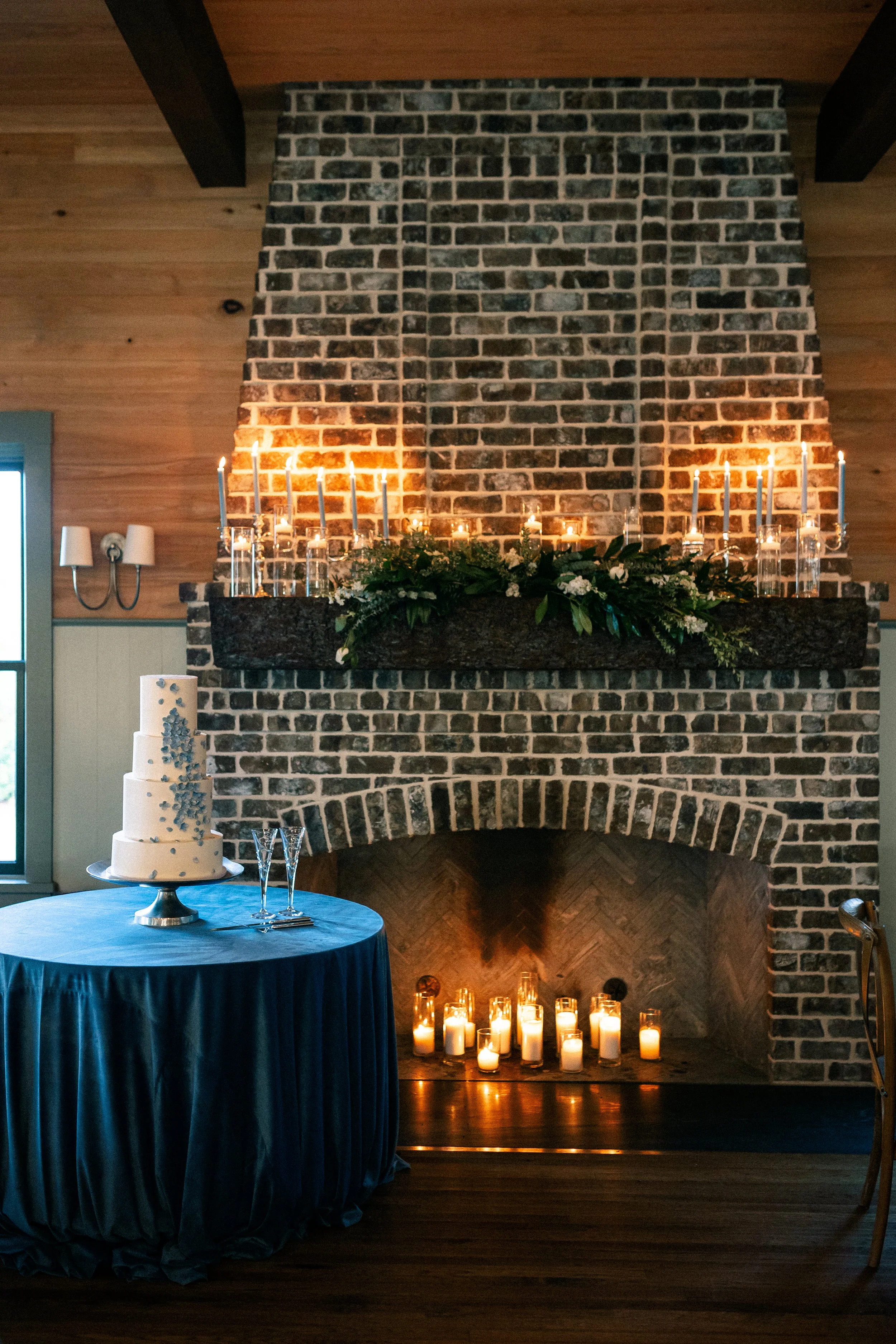 A fireplace decorated with candles and greenery, with a white cake and champagne glasses on a table nearby, suggesting a festive or romantic setting.