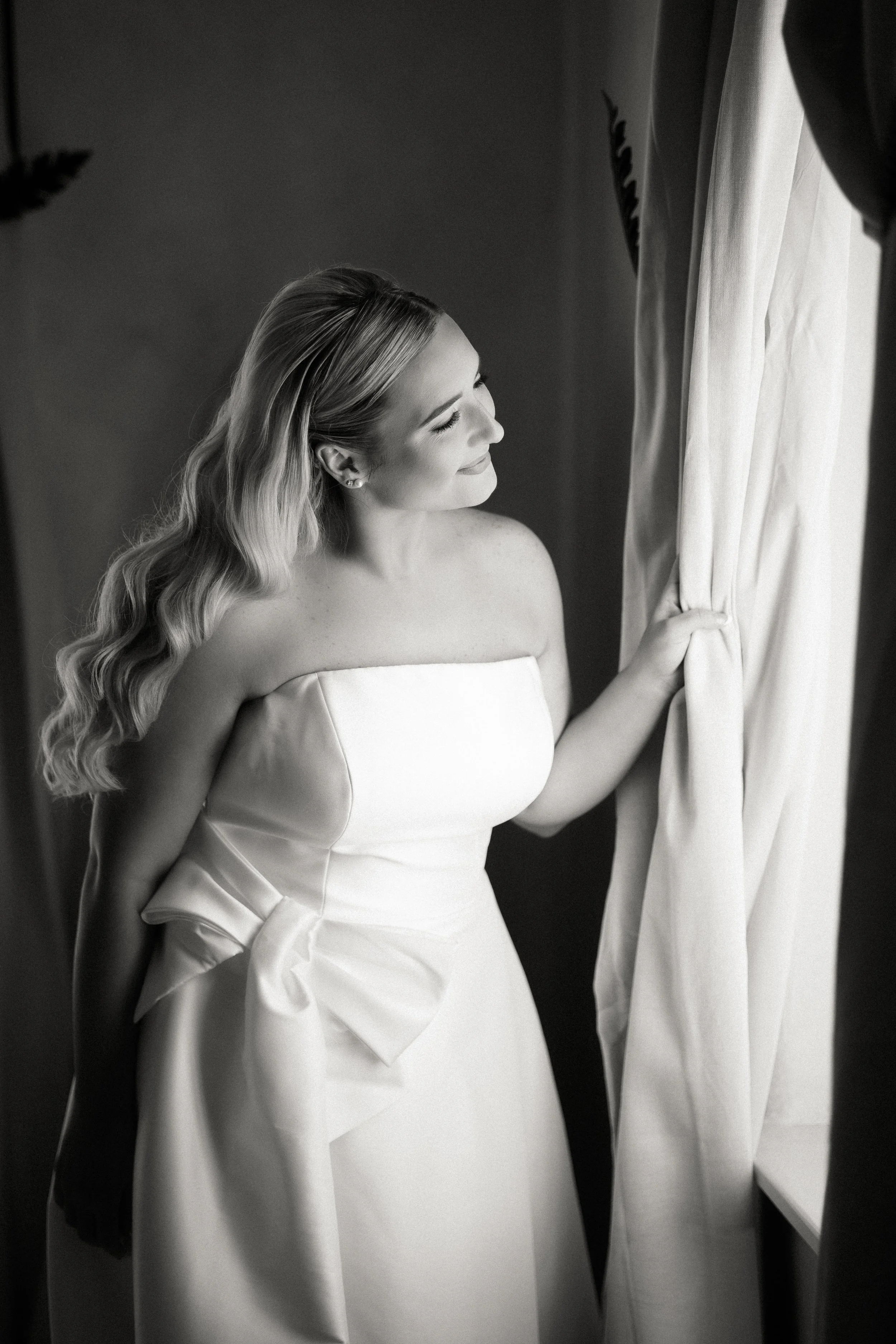 Black and white photo of a woman in a strapless wedding dress with a bow, smiling gently as she looks out a window and gently pulls back a curtain.