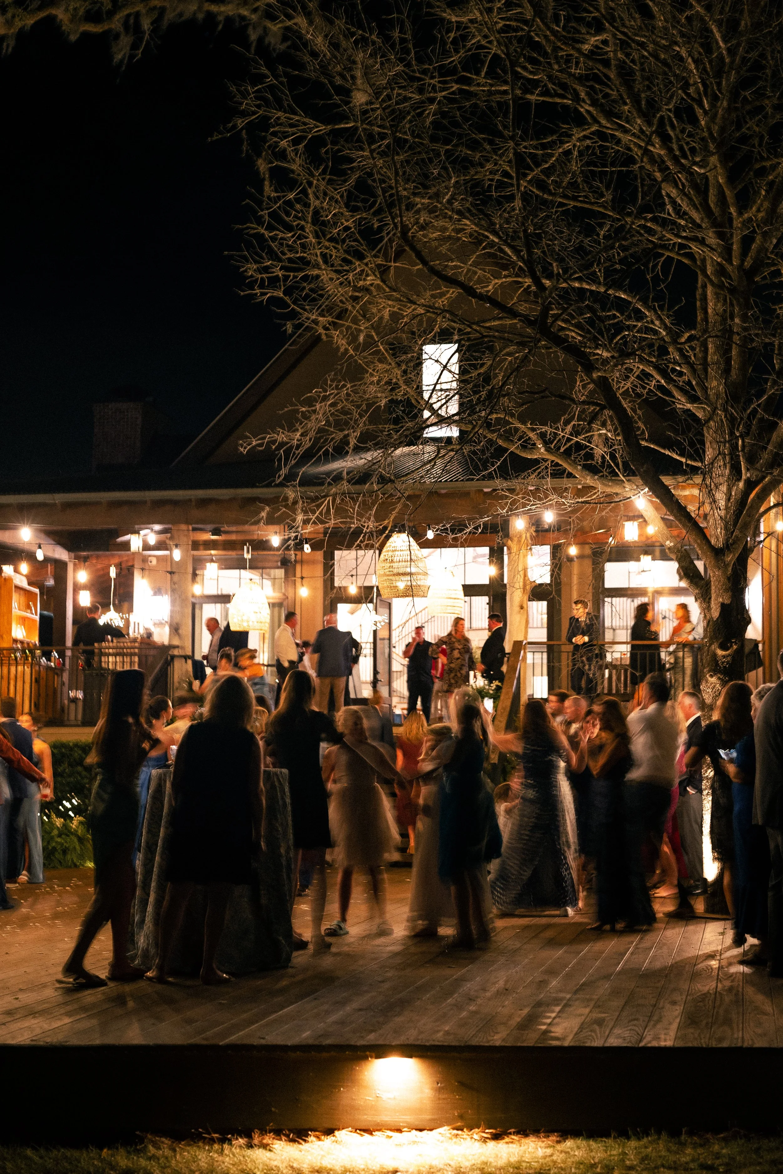 People dancing and socializing at an outdoor night party on a wooden deck, illuminated by string lights and hanging lamps, with a house or pavilion in the background and a large tree in the foreground.