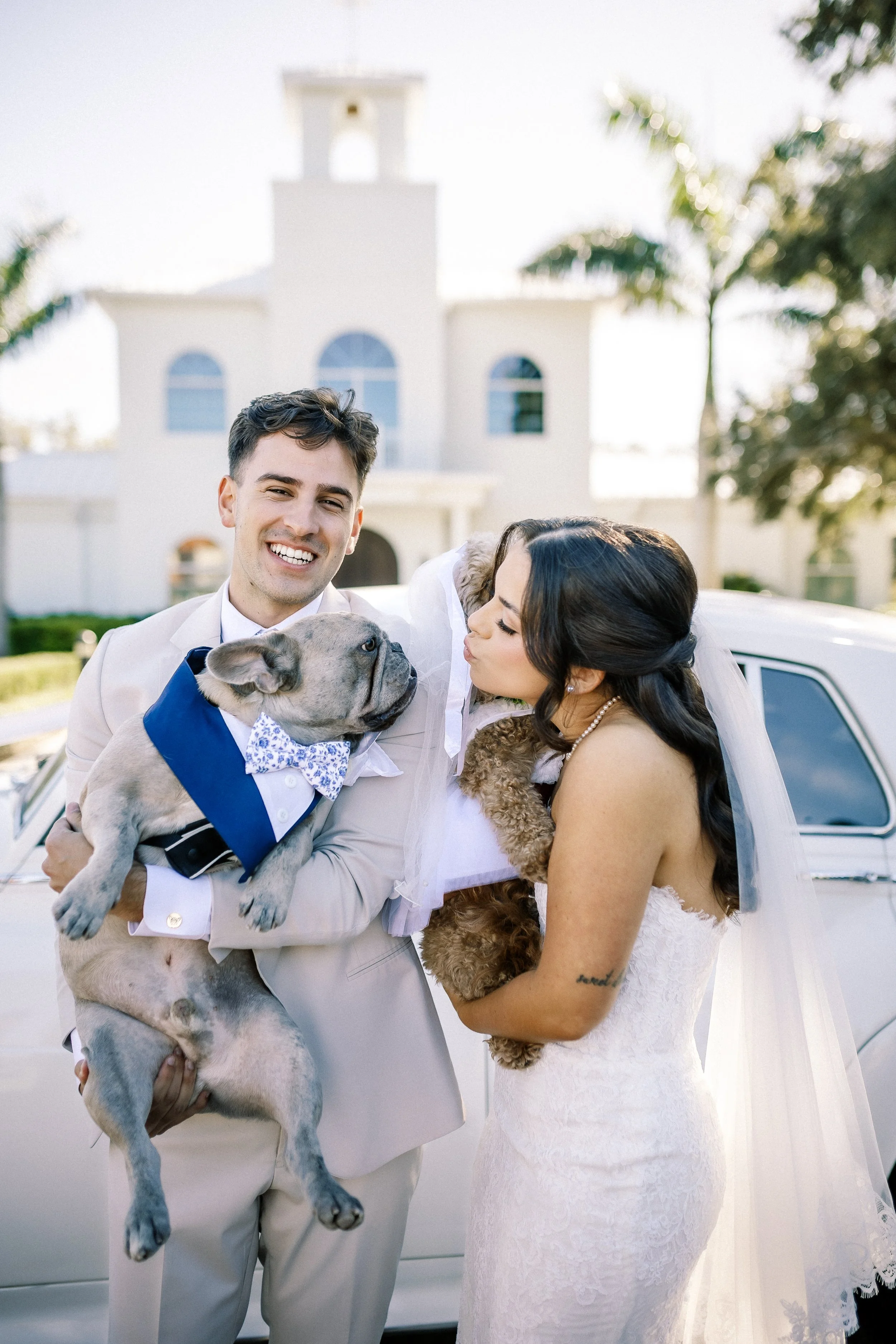 A happy bride and groom holding their dogs, a French Bulldog and a poodle, at their wedding outside in front of a church, with a vintage car in the background.