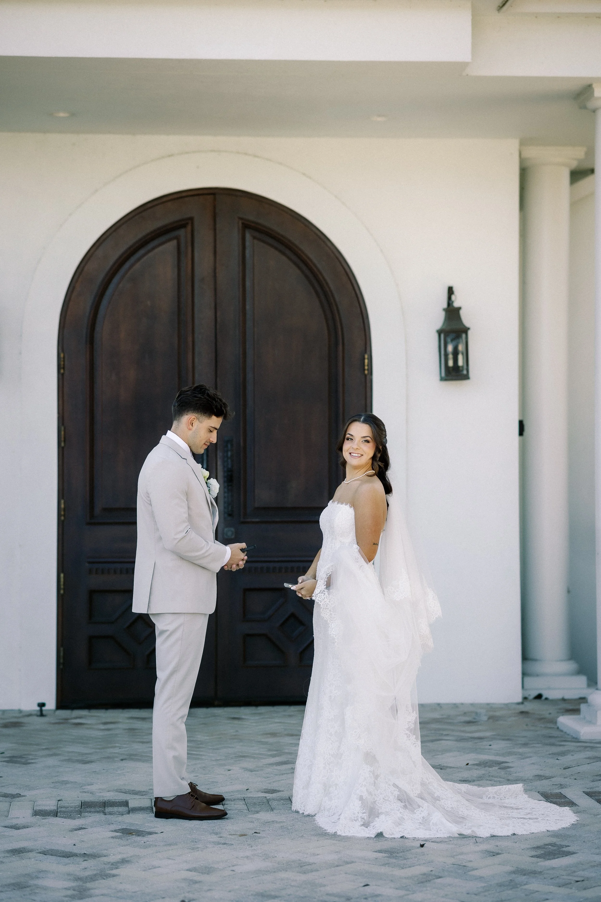 A bride and groom standing outside a building with a dark wooden door and white walls. The bride is smiling at the camera, wearing a white lace wedding gown with a train, and the groom is looking at his phone, dressed in a light-colored suit.