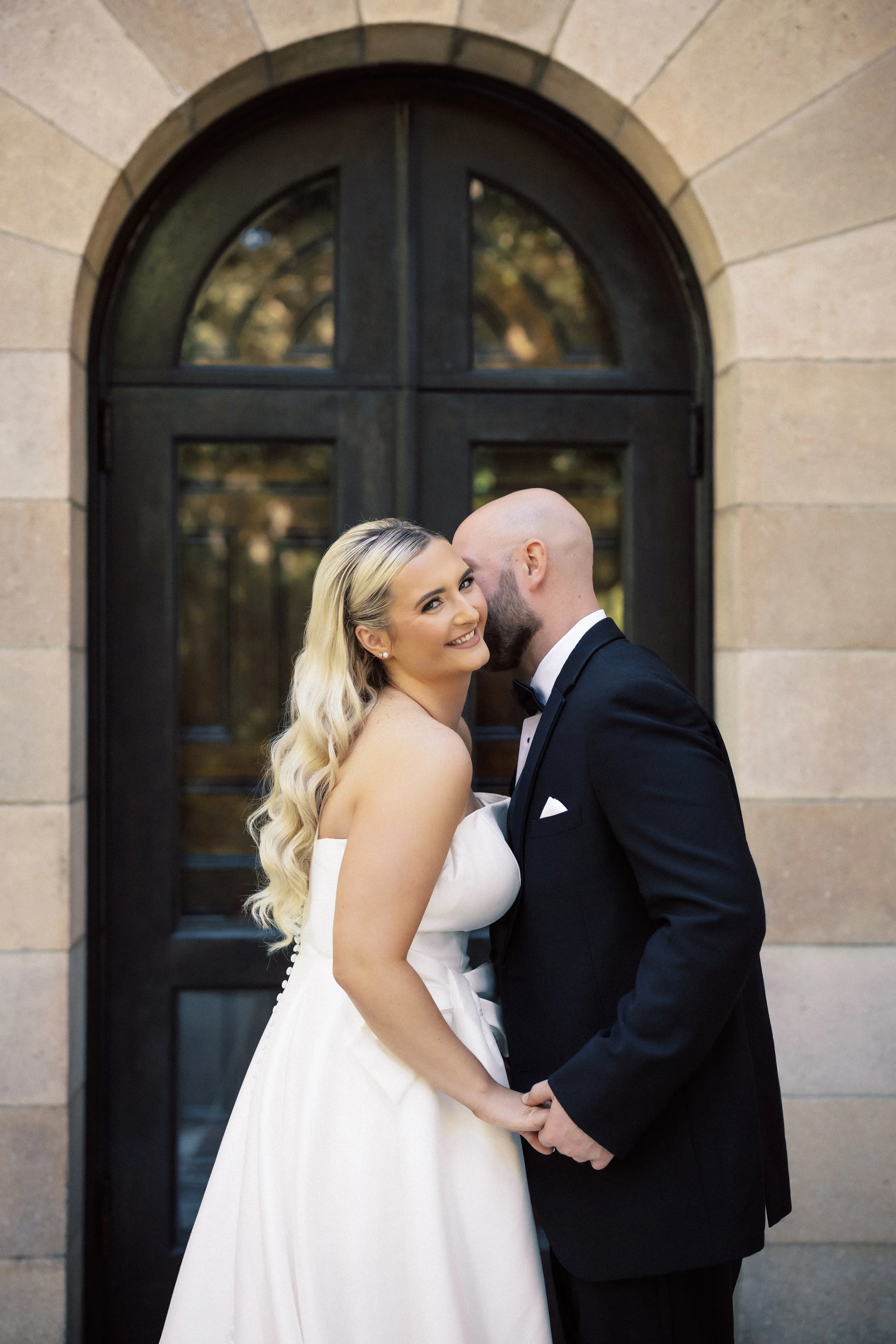 Bride and groom holding hands and sharing a kiss in front of a large, arched window.