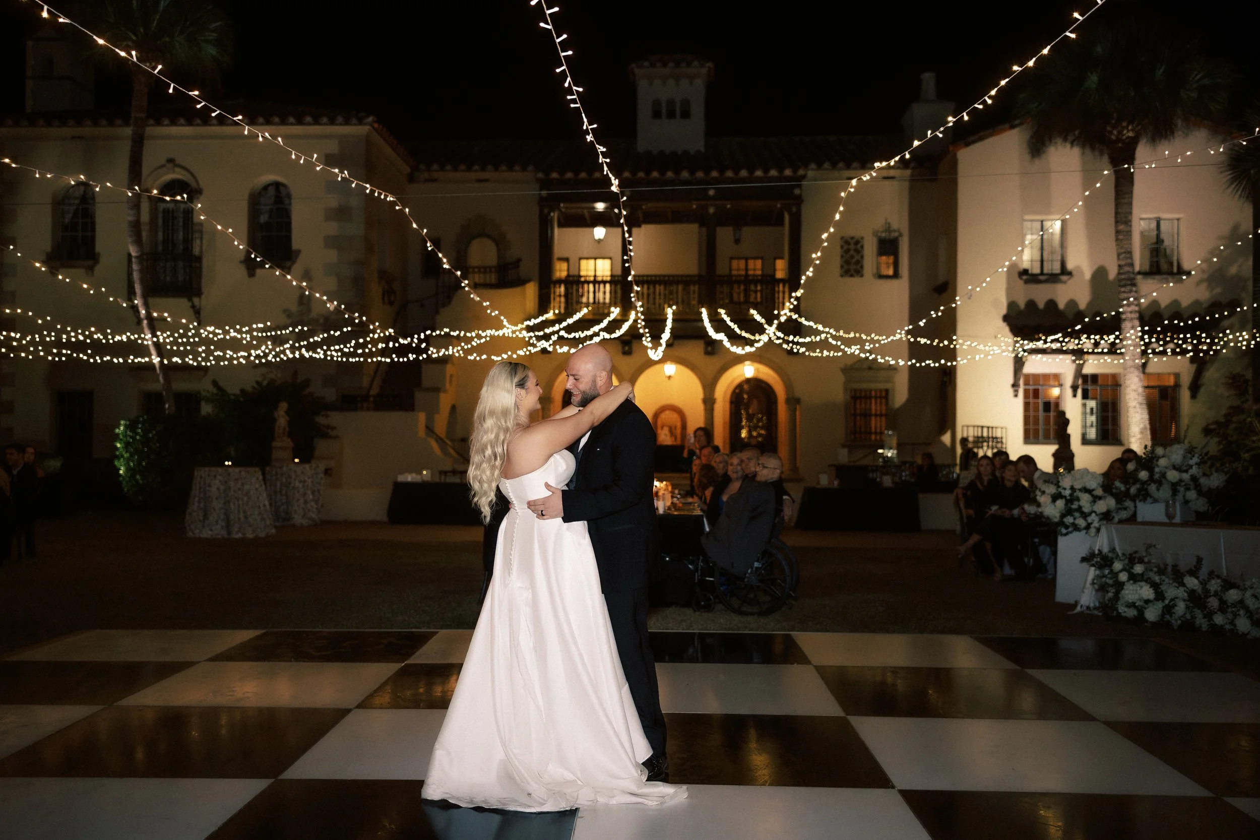 Couple dancing at their wedding reception outdoors at night under string lights, with guests seated around and a large mansion-style building in the background.