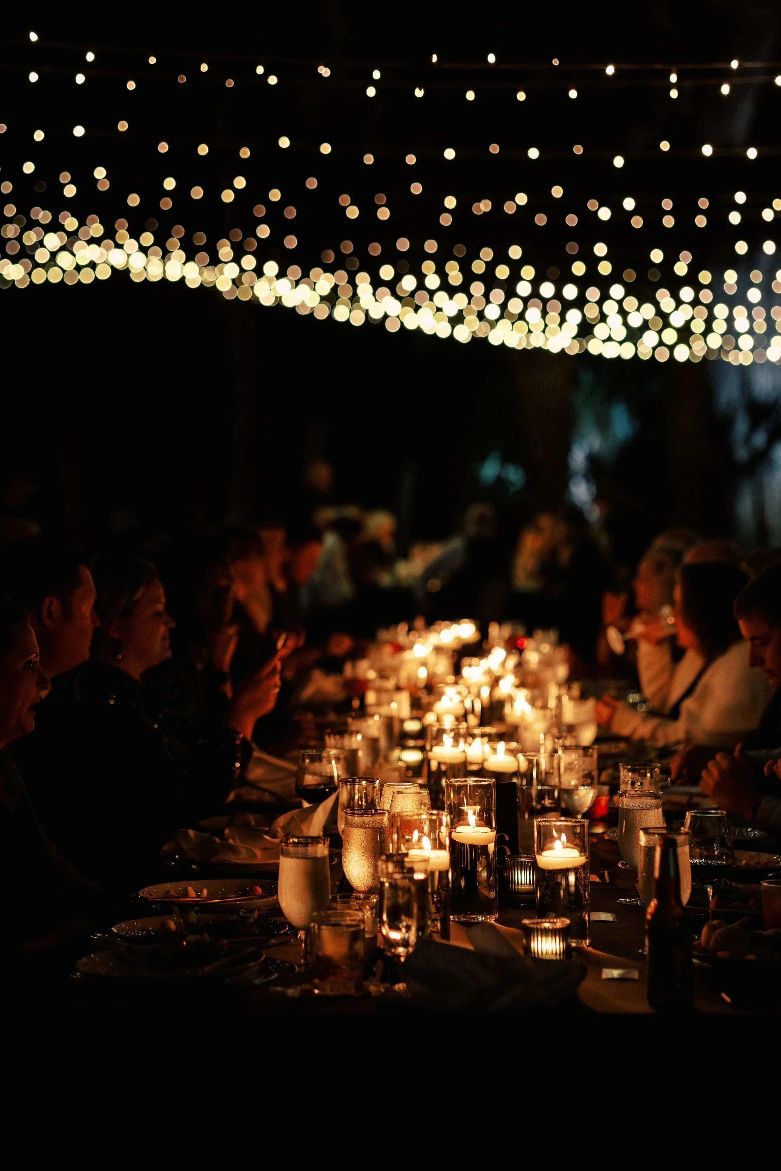 People sitting at a long dinner table illuminated by candles during an outdoor event at night, with string lights overhead creating a bokeh effect.