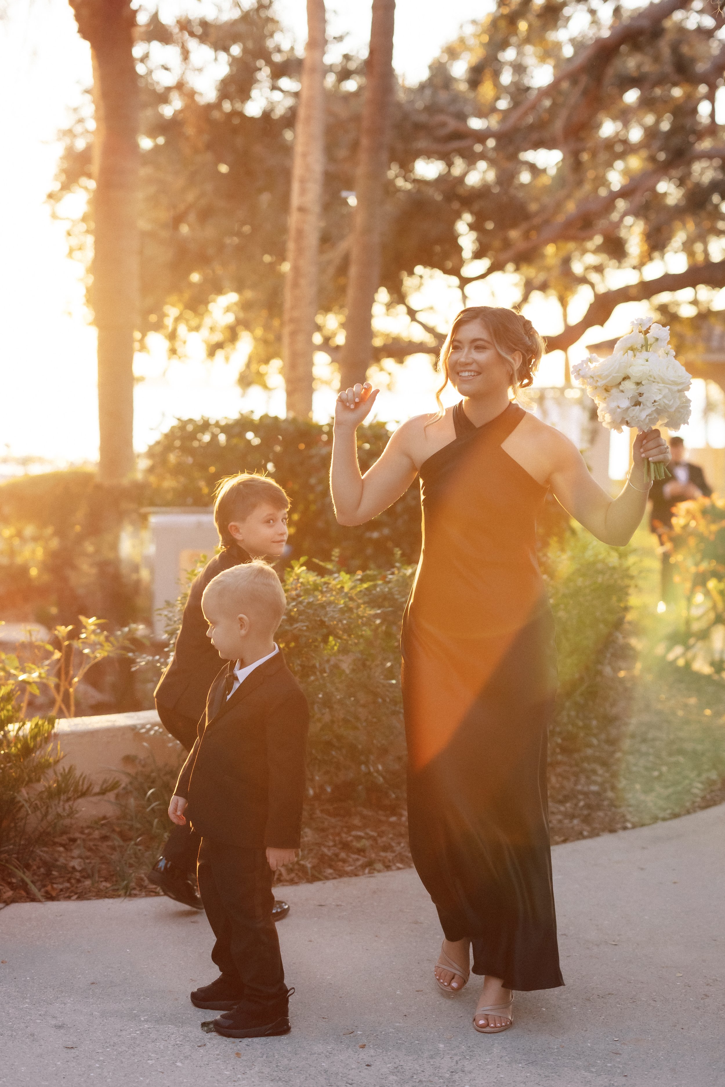 A woman in a black evening gown holding a bouquet of white flowers smiling joyfully as she walks outdoors during sunset, flanked by two young boys in black suits. Trees and foliage are visible in the background.