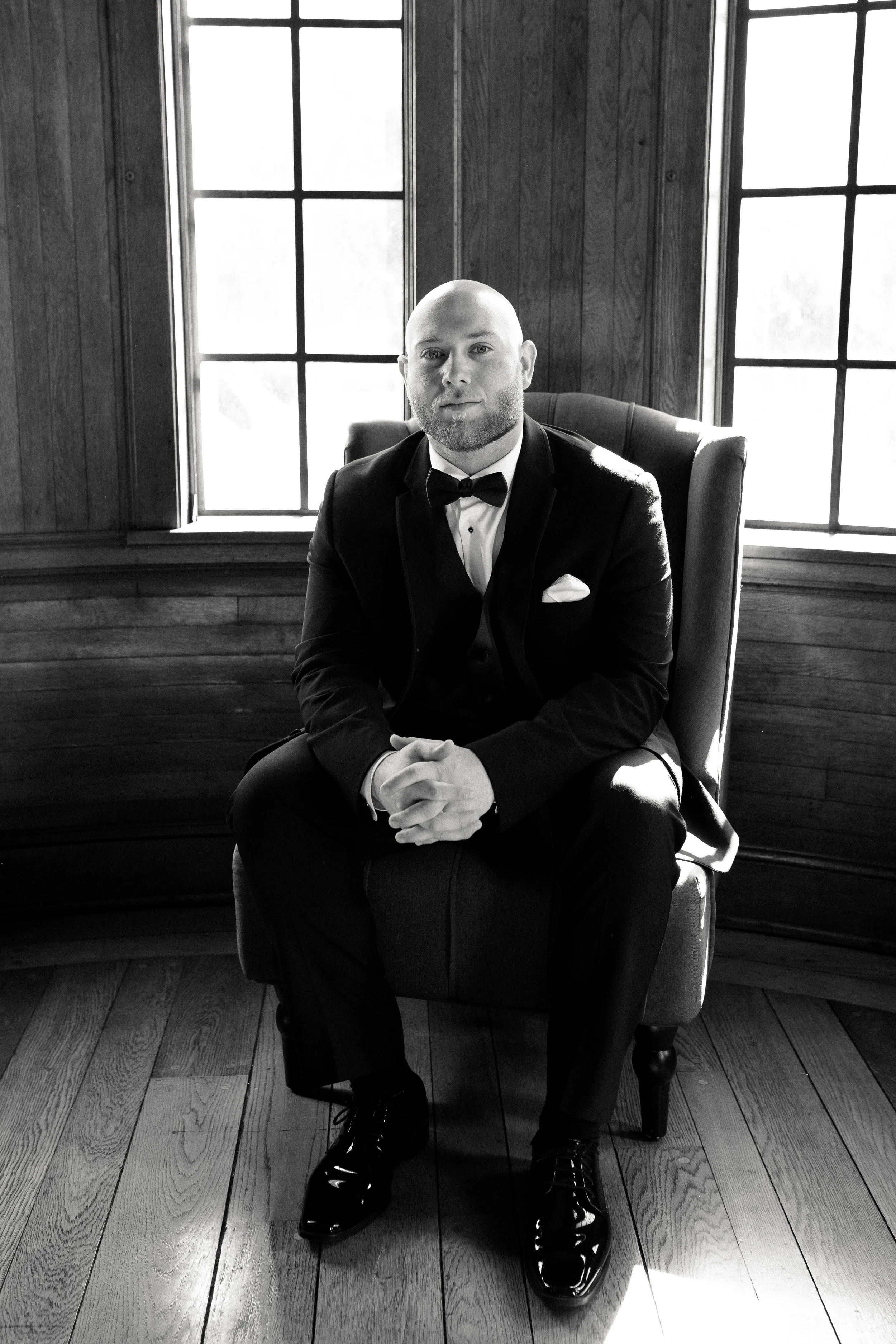 A man in a tuxedo sitting with hands clasped on an armchair in front of windows with wooden frames, black and white photo.