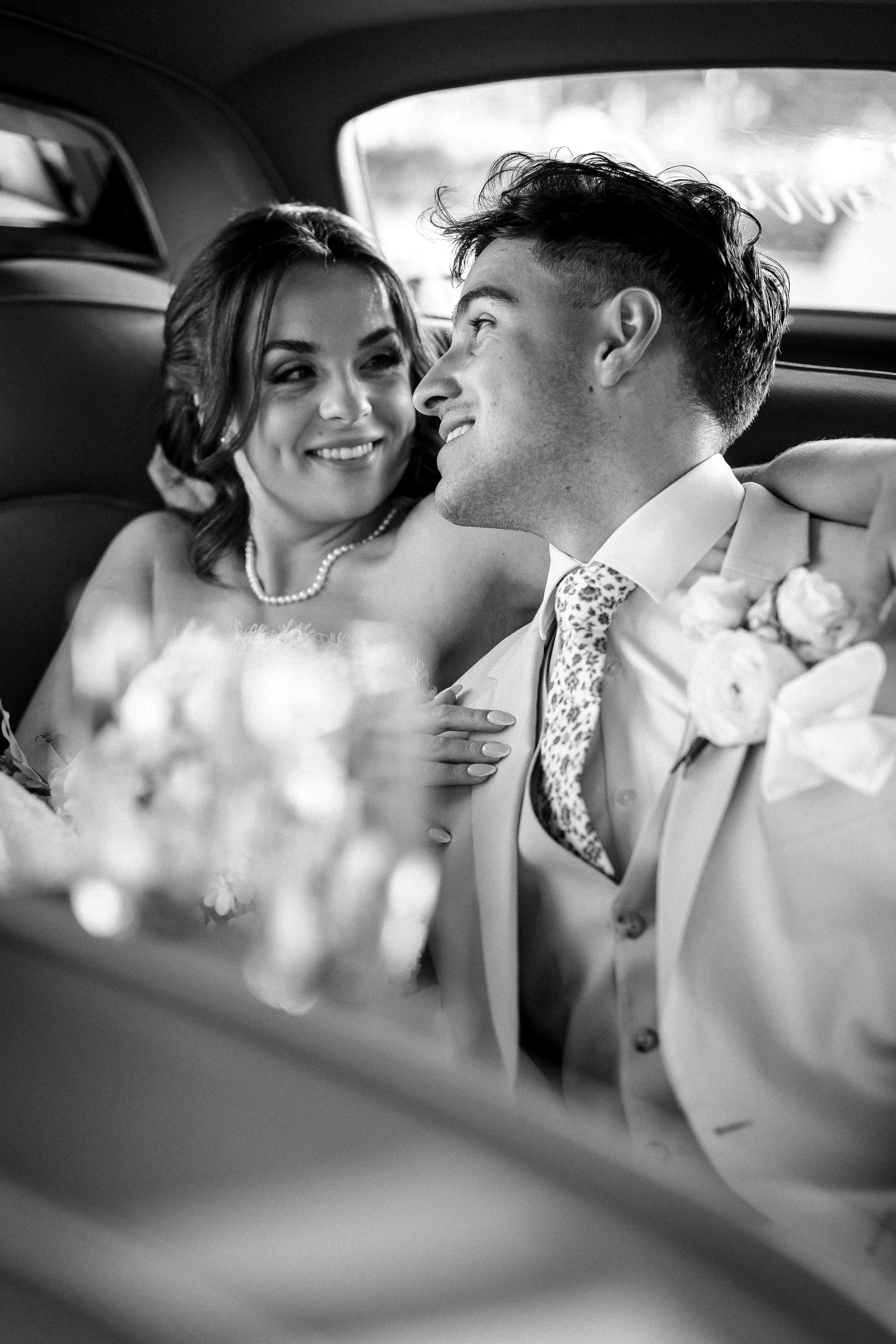 Black and white photo of a wedding couple sitting in a car, smiling at each other.