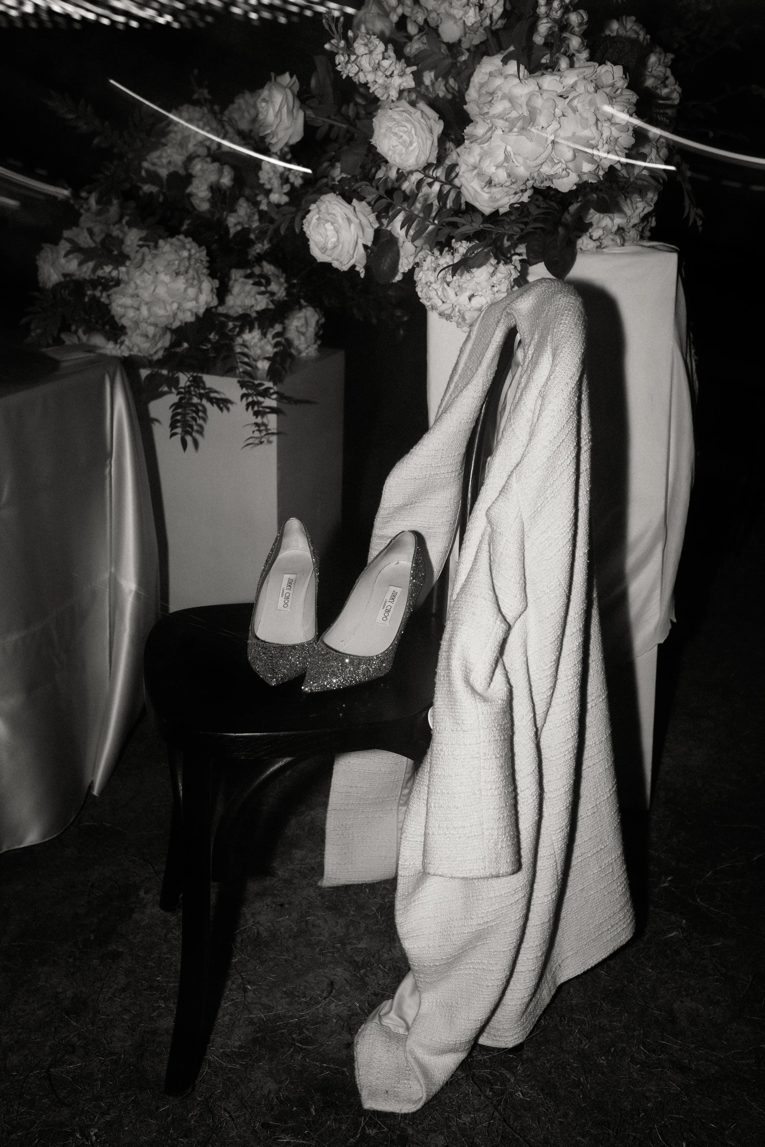 A pair of sparkling high heel shoes on a black chair, with a light-colored coat draped over the back, next to a floral arrangement of roses and hydrangeas at an event or wedding reception.