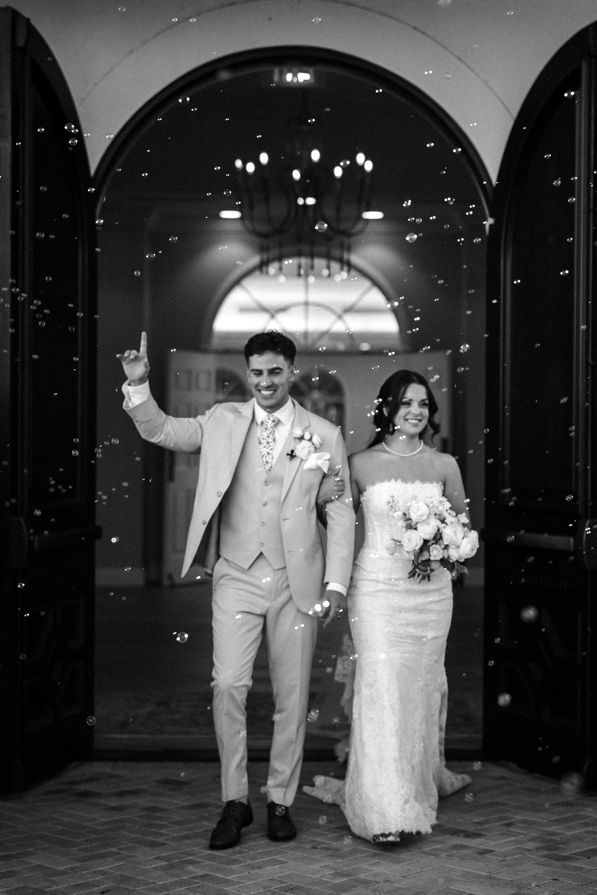 A bride and groom entering a venue through open doors, smiling and celebrating with bubbles. The groom is raising one finger in the air, and the bride is holding a bouquet of flowers.