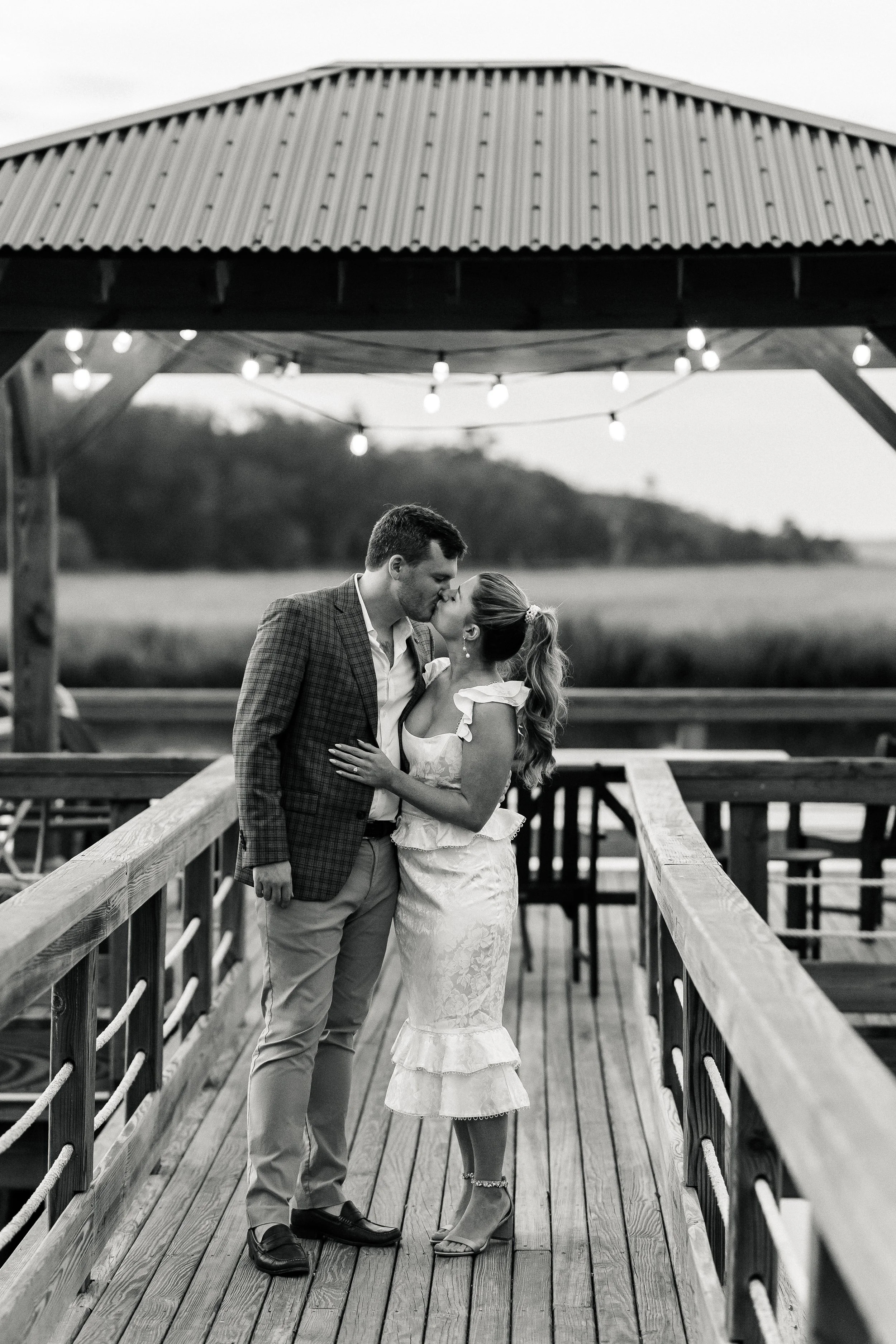 A black and white photo of a couple kissing on a wooden dock under a pavilion with string lights, with a field and trees in the background.