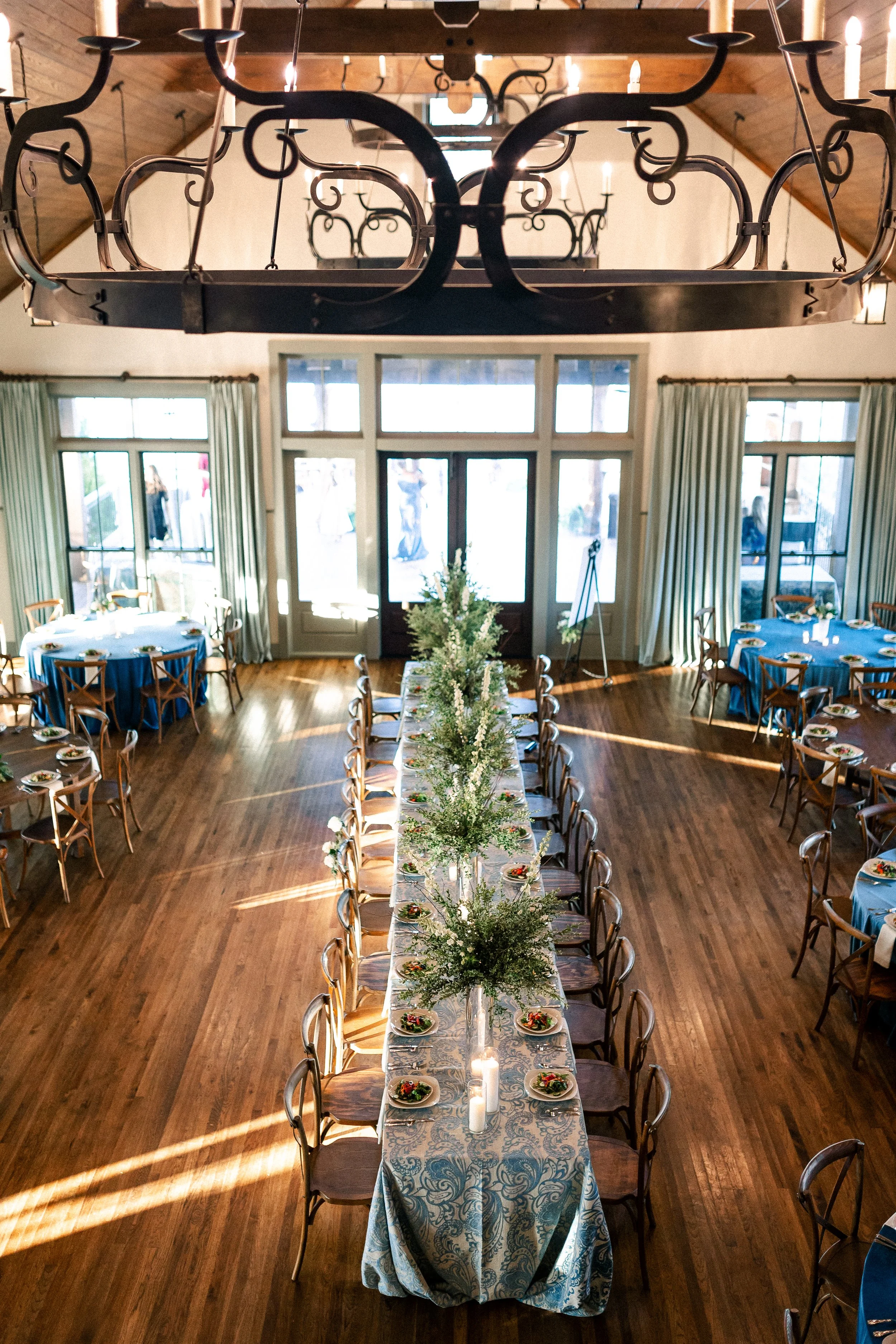 Wedding reception hall with a long central table decorated with floral arrangements and candles, surrounded by chairs, with additional round tables set for guests, large windows with curtains, and a large wrought iron chandelier hanging from a wooden
