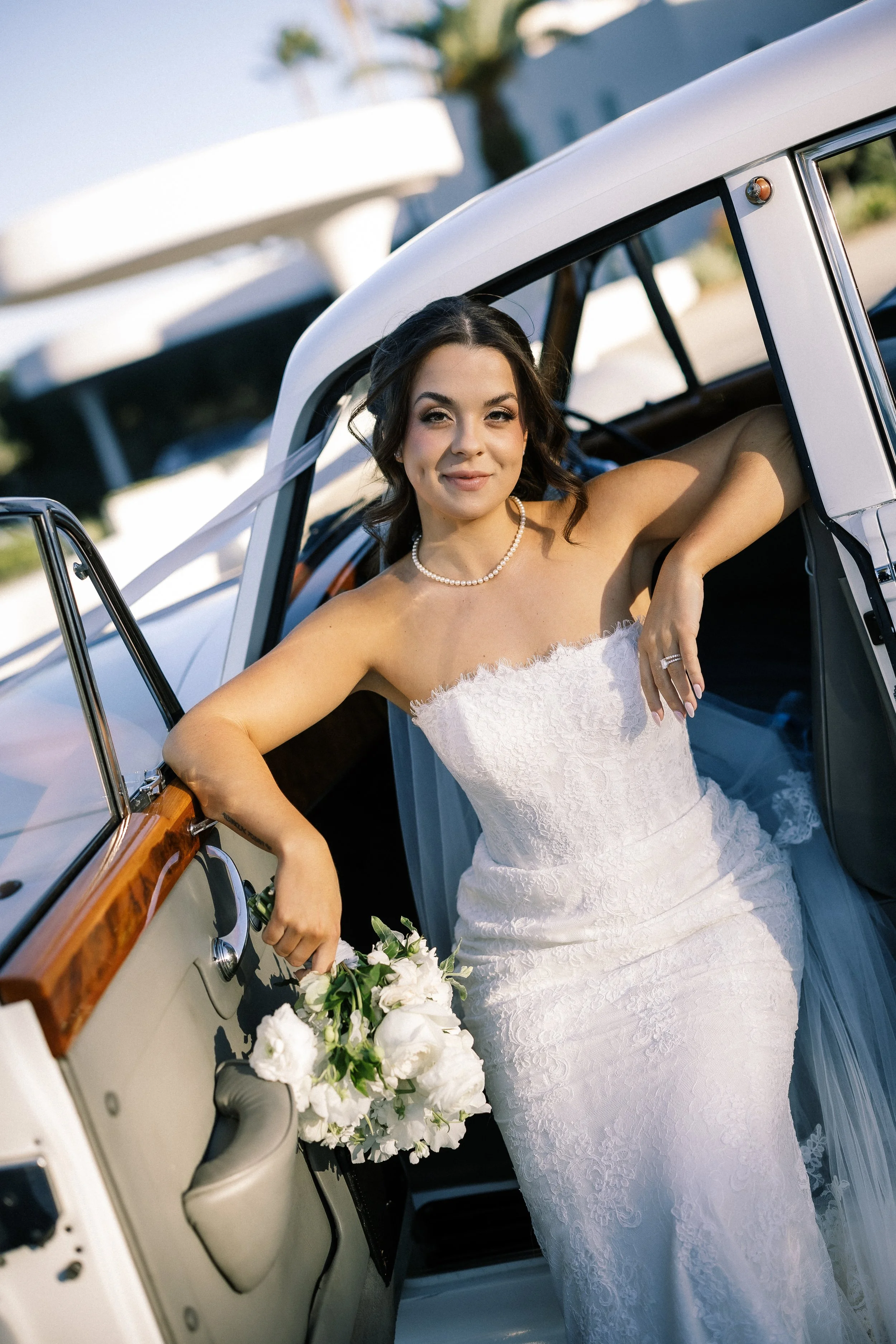 A bride in a white strapless wedding dress with lace details, holding a bouquet of white flowers, leaning on the door of a vintage white car with wooden interior trim, smiling and looking at the camera as she poses outdoors on a sunny day.