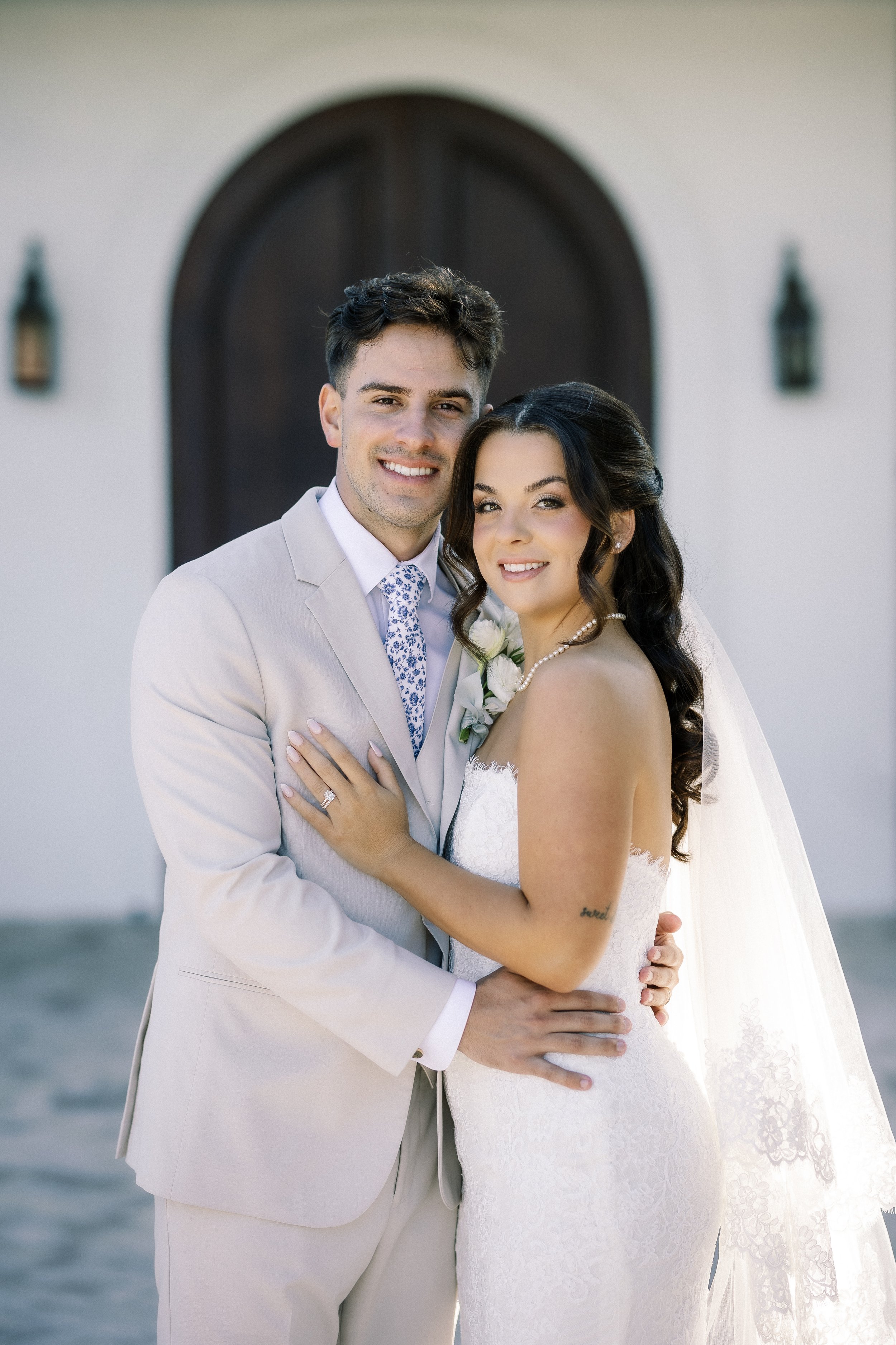 A newlywed couple in wedding attire standing close together outdoors in front of a building with an arched dark wooden door and wall lanterns, smiling at the camera.