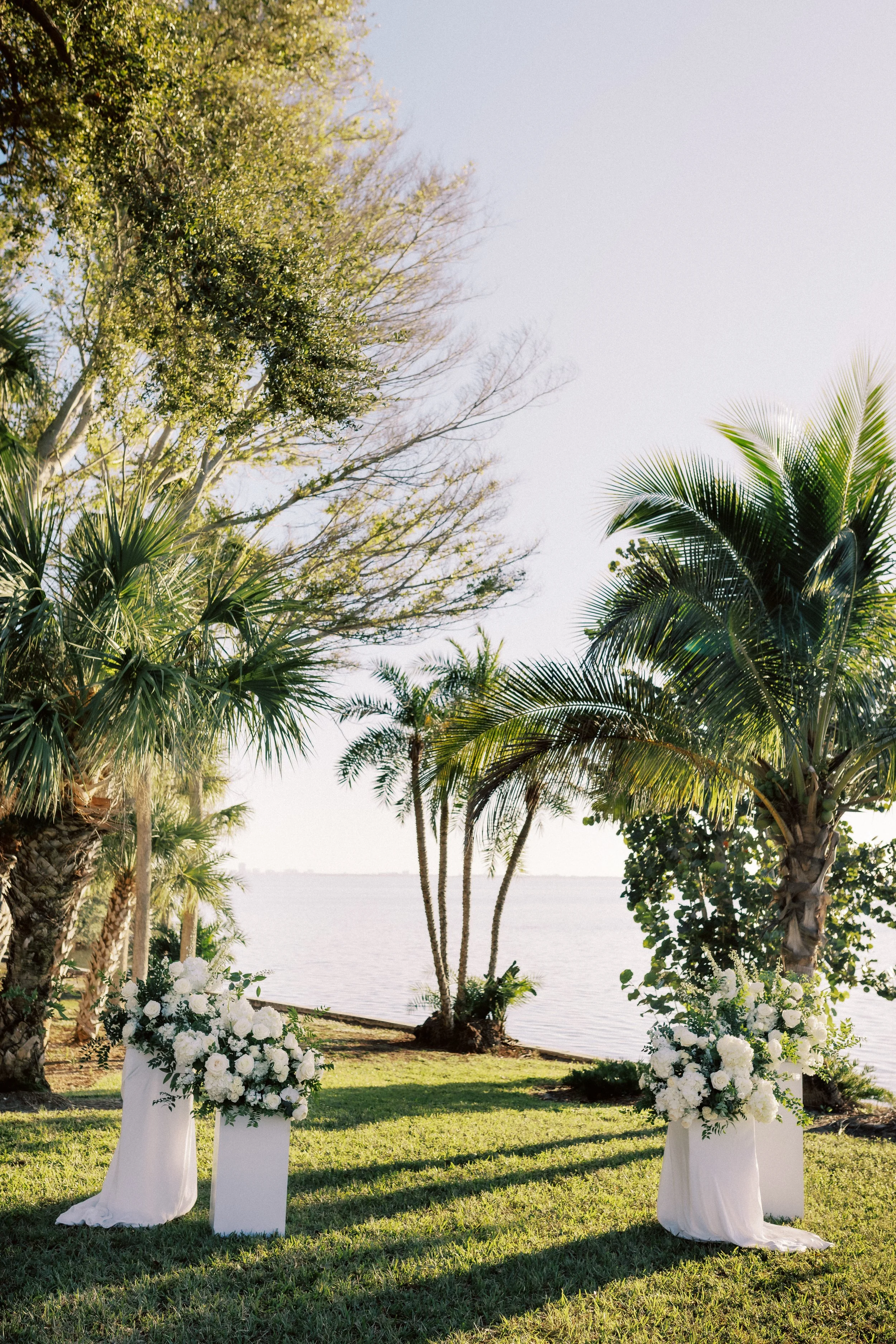 Decorated outdoor wedding ceremony altar with white floral arrangements and white drapes, overlooking a body of water with palm trees and other greenery.