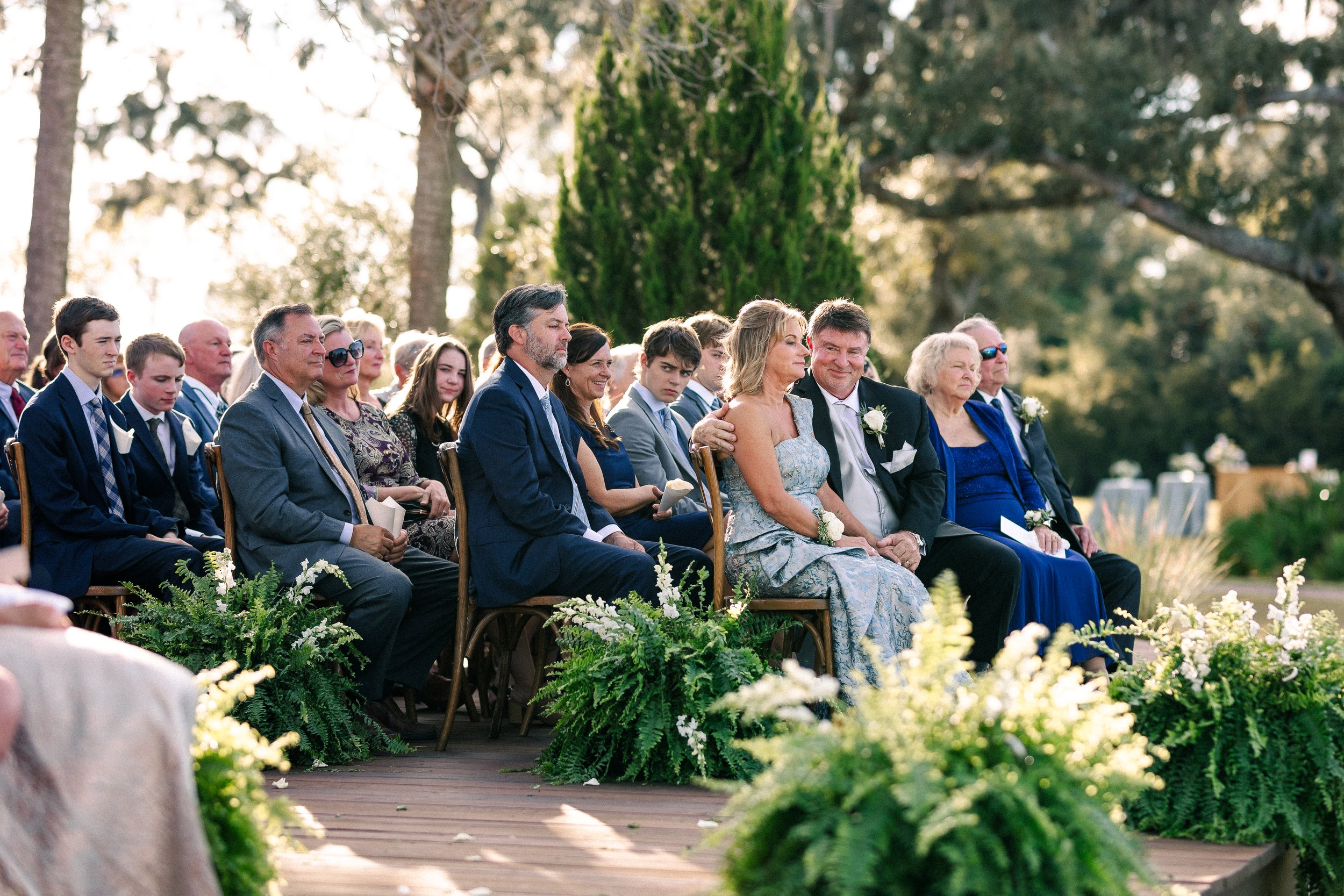 People seated outdoors during a wedding ceremony, with trees and greenery in the background, and floral decorations in the foreground.