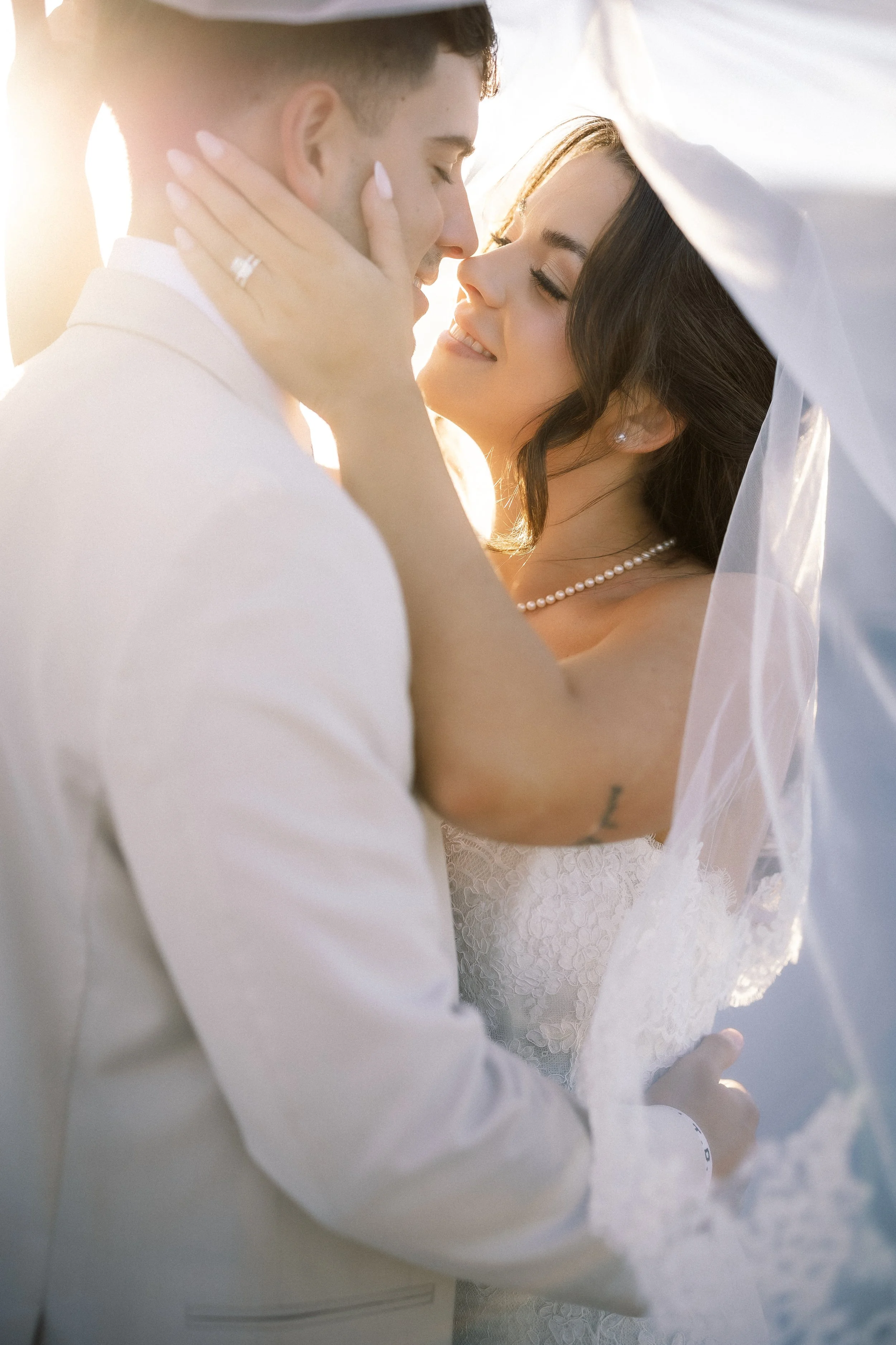 A bride and groom sharing a close, intimate moment under a wedding veil, with the bride smiling and the groom gently touching her face, during sunset.