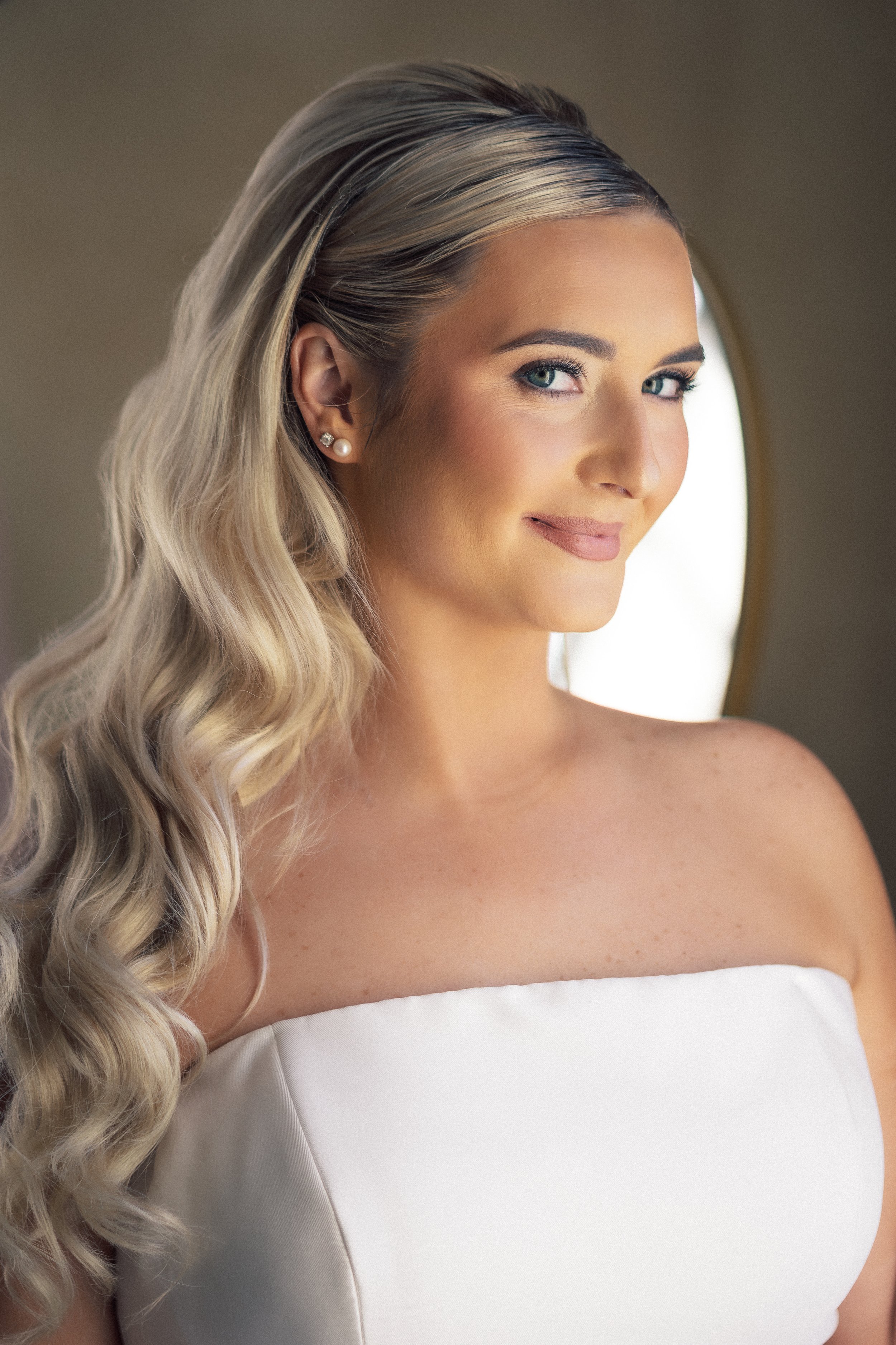 A woman with long, wavy blonde hair and blue eyes, wearing pearl earrings, smiles softly in front of a mirror.