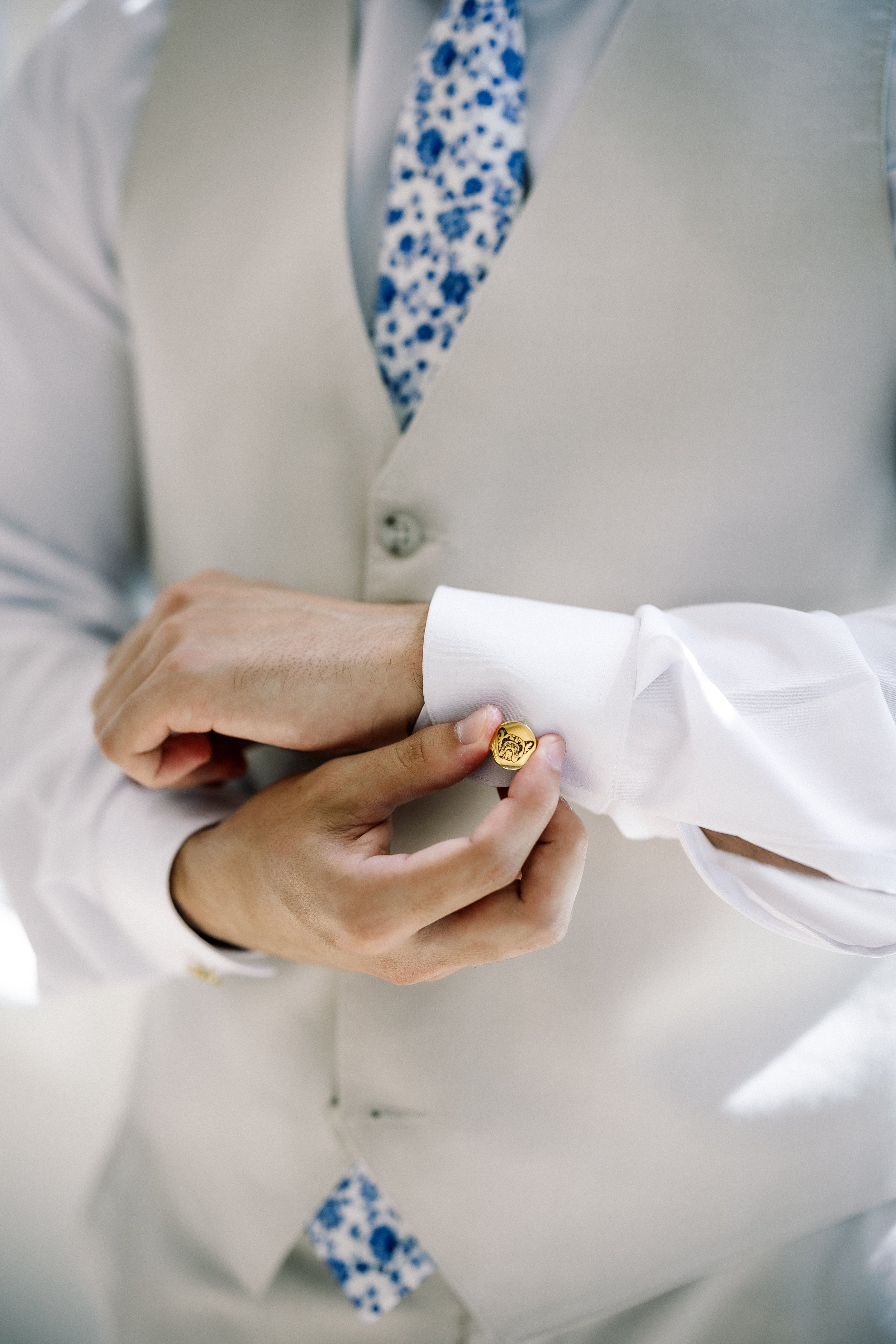Person adjusting a gold cufflink on a white shirt cuff, wearing a white vest and a patterned blue tie.