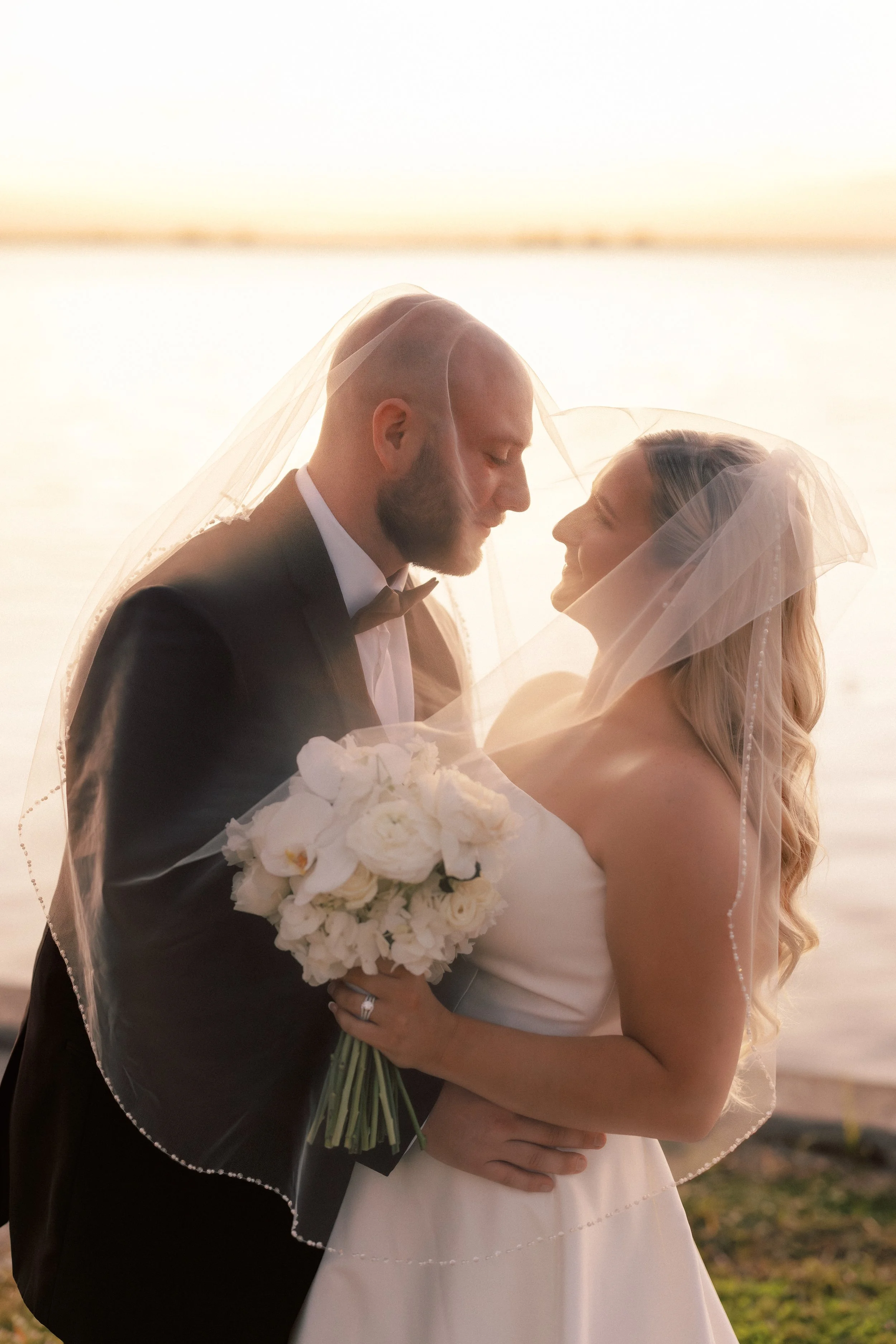 A wedding couple stands close by a body of water at sunset, sharing an intimate moment under a sheer veil, with the bride holding a bouquet of white flowers.