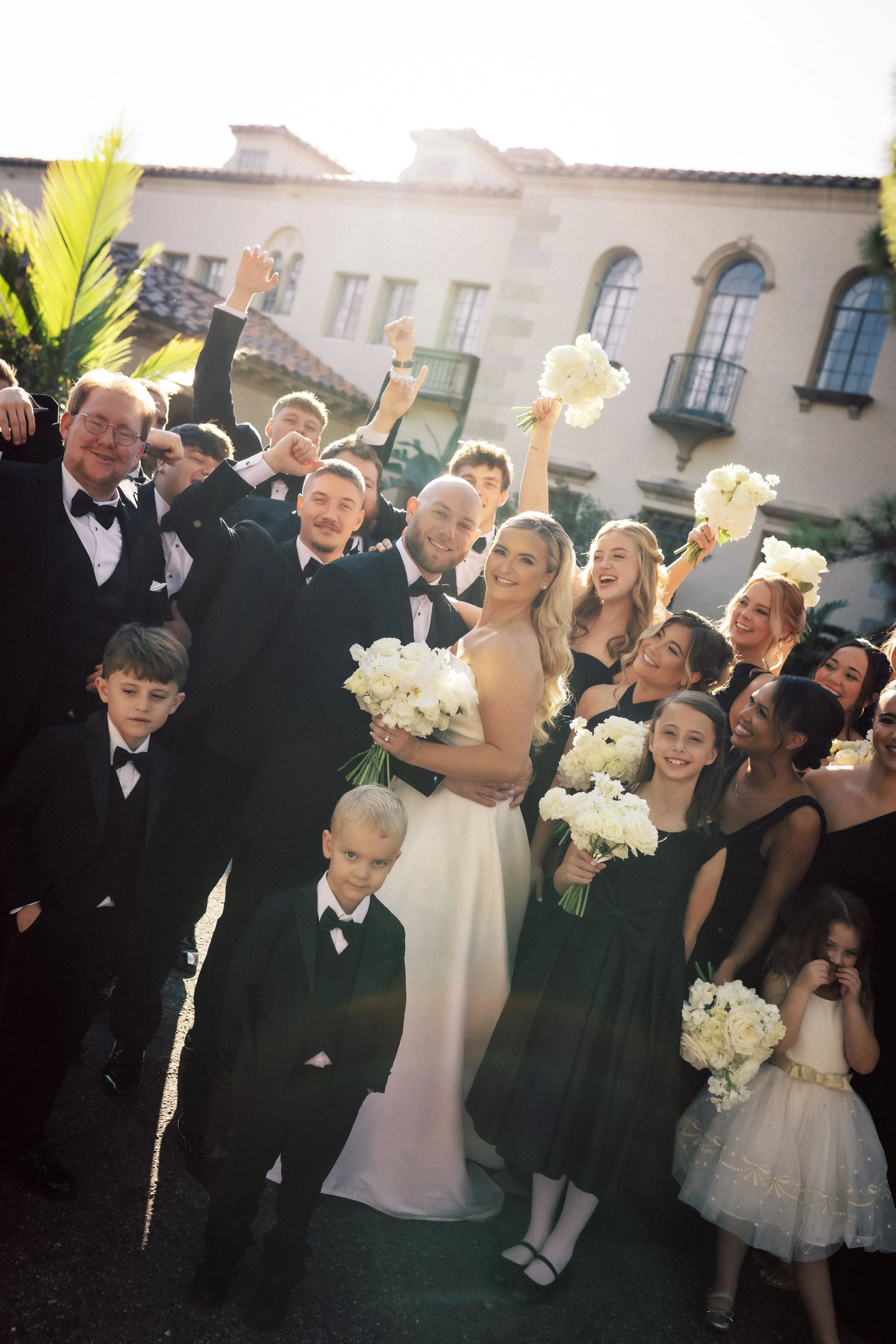 Group of people at a wedding celebration wearing formal attire, with the bride in a white gown holding a bouquet, and others holding flowers, smiling and celebrating outdoors in front of a large building.