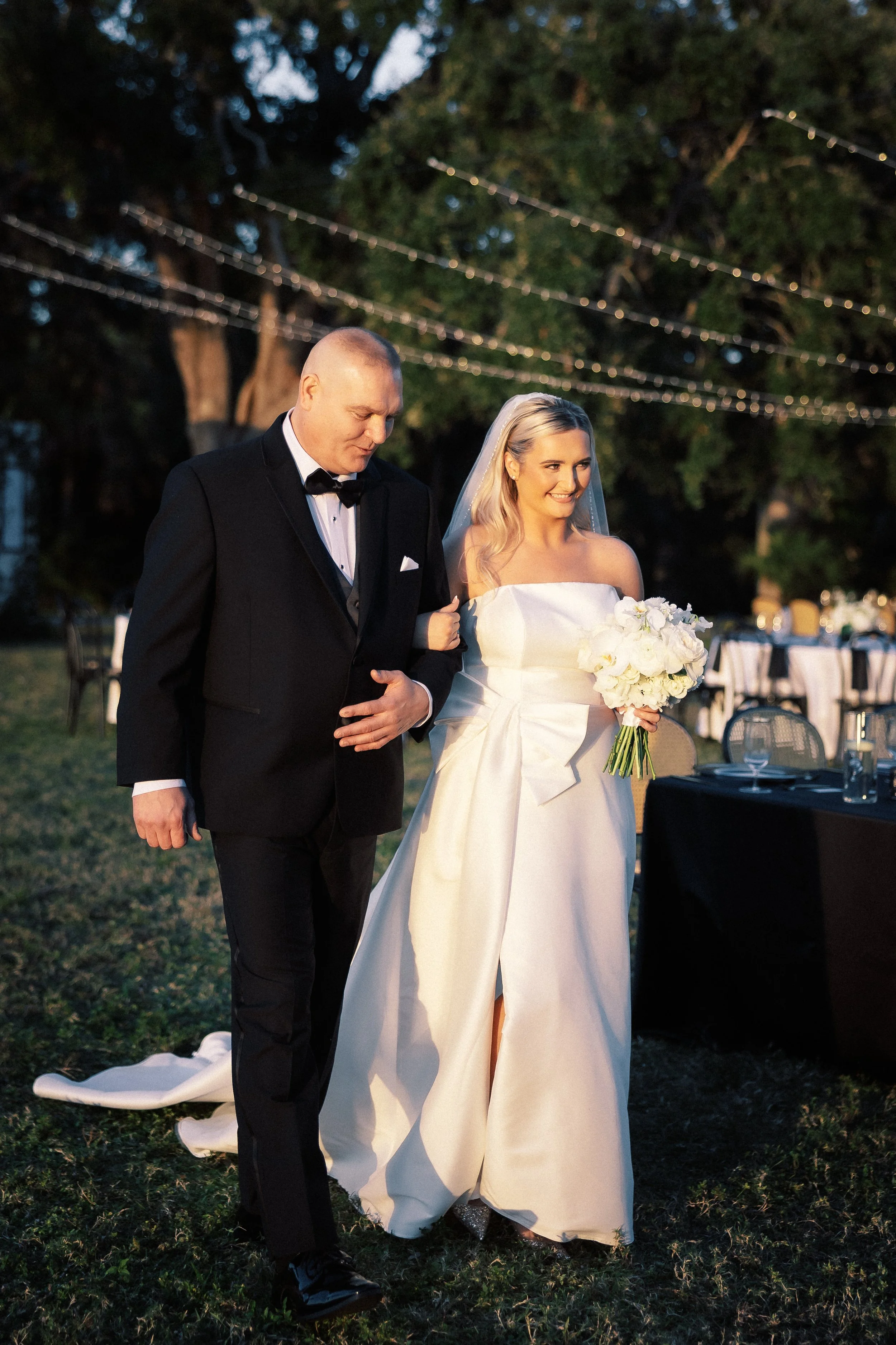 A bride with a bouquet walking with a man in a tuxedo outdoors during her wedding at sunset.