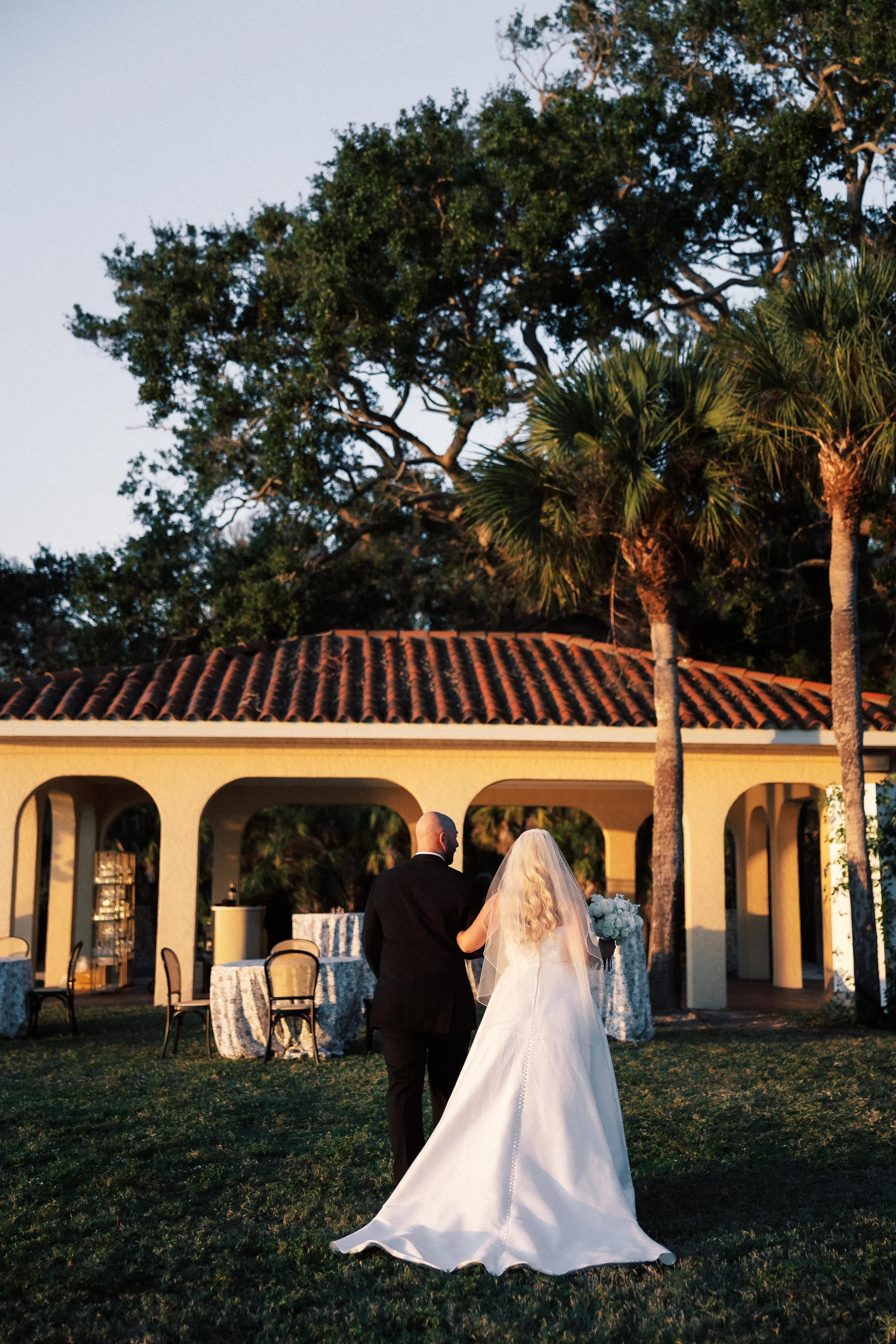 A bride and groom walk together outdoors during sunset at a wedding, with palm trees, a building with arches, and decorated tables in the background.