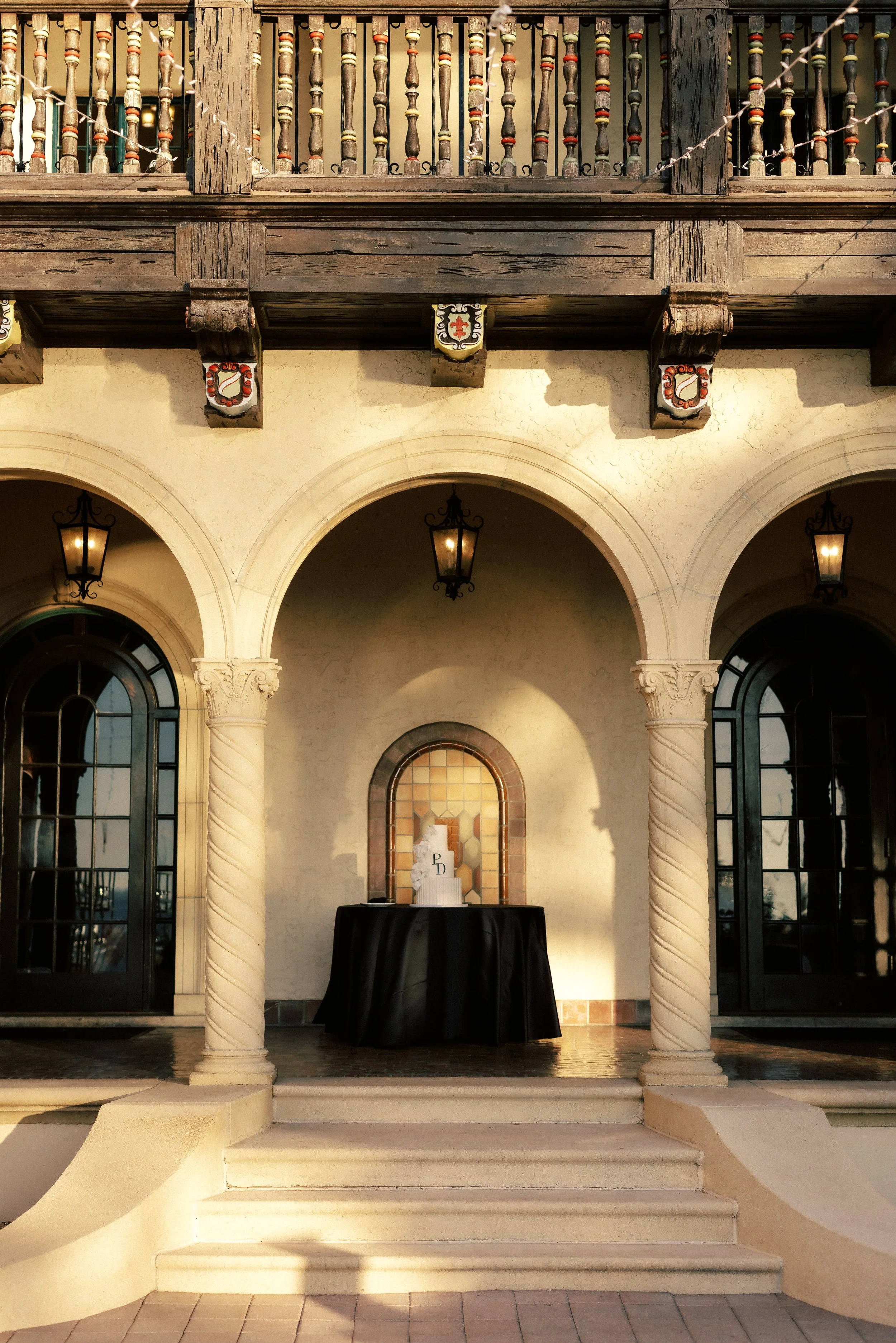 Exterior of a Mediterranean-style building with arched doorways, decorative columns, and a balcony with a wooden railing. A small table with a black tablecloth and a decorative backdrop is positioned under one of the arches, illuminated by warm light