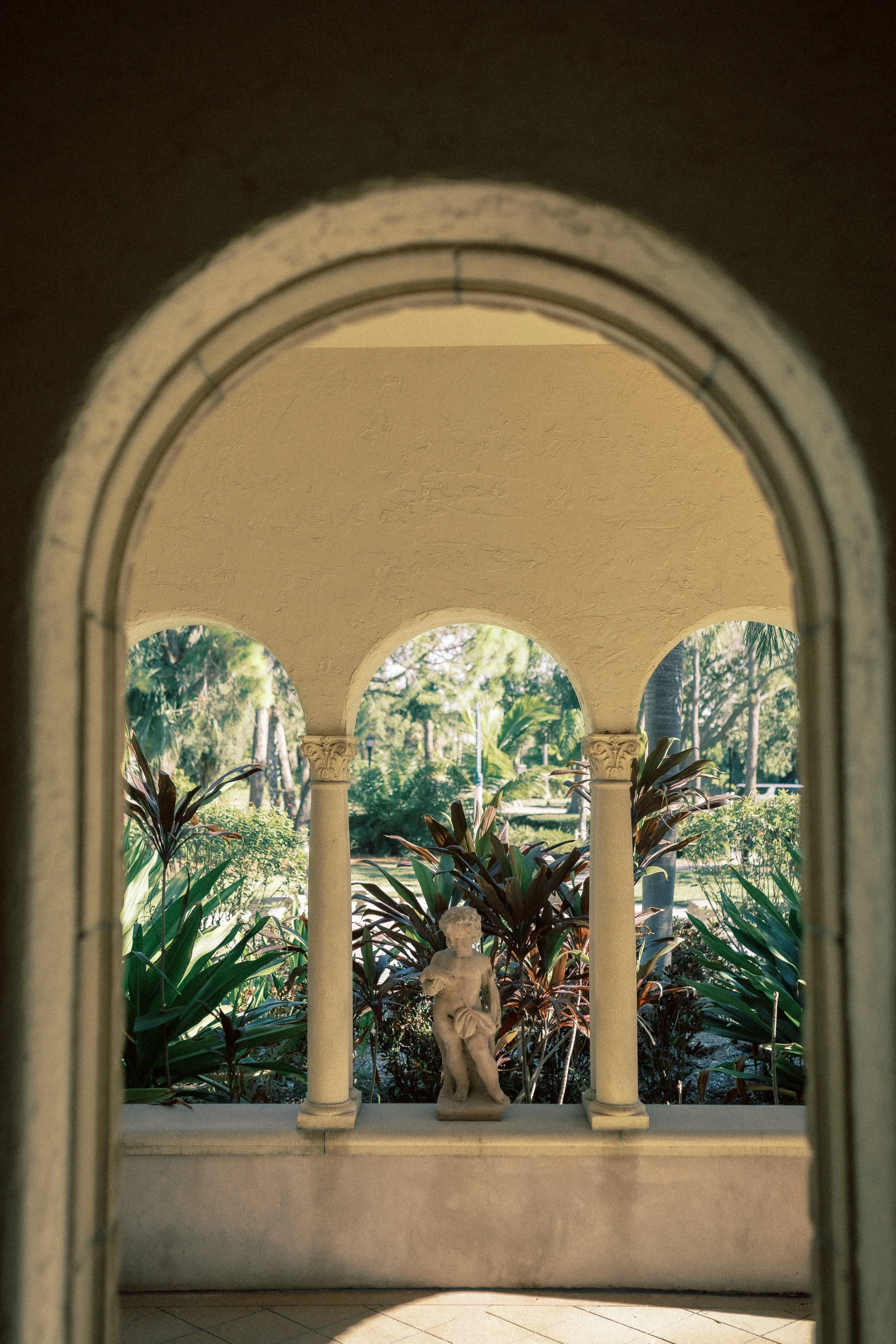 View through an arched window showing a garden with a statue of a child, lush plants, and trees outside.