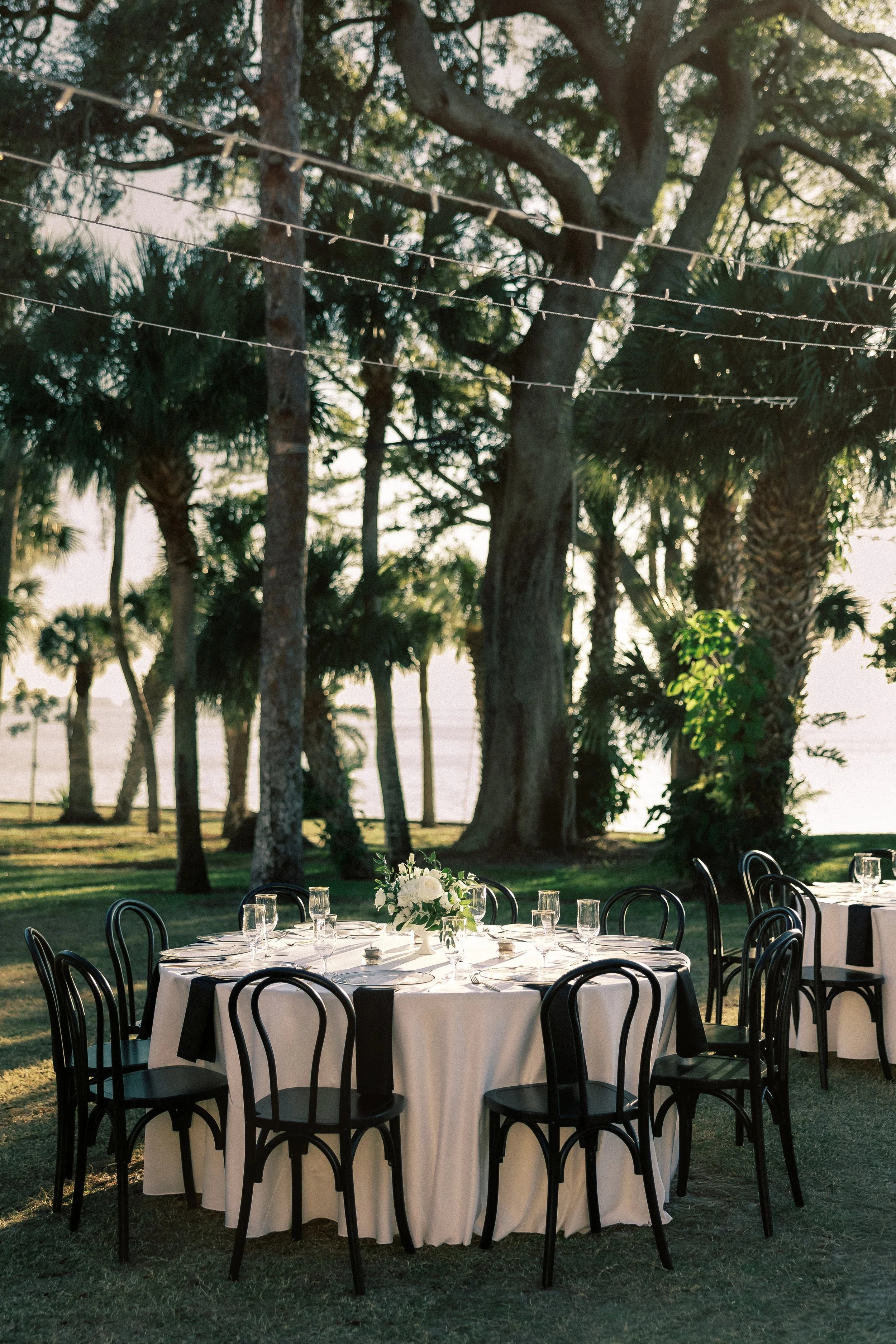 Outdoor wedding or event setup with round table, white tablecloth, black chairs, floral centerpiece, and string lights in a lush, wooded area near a body of water at sunset.