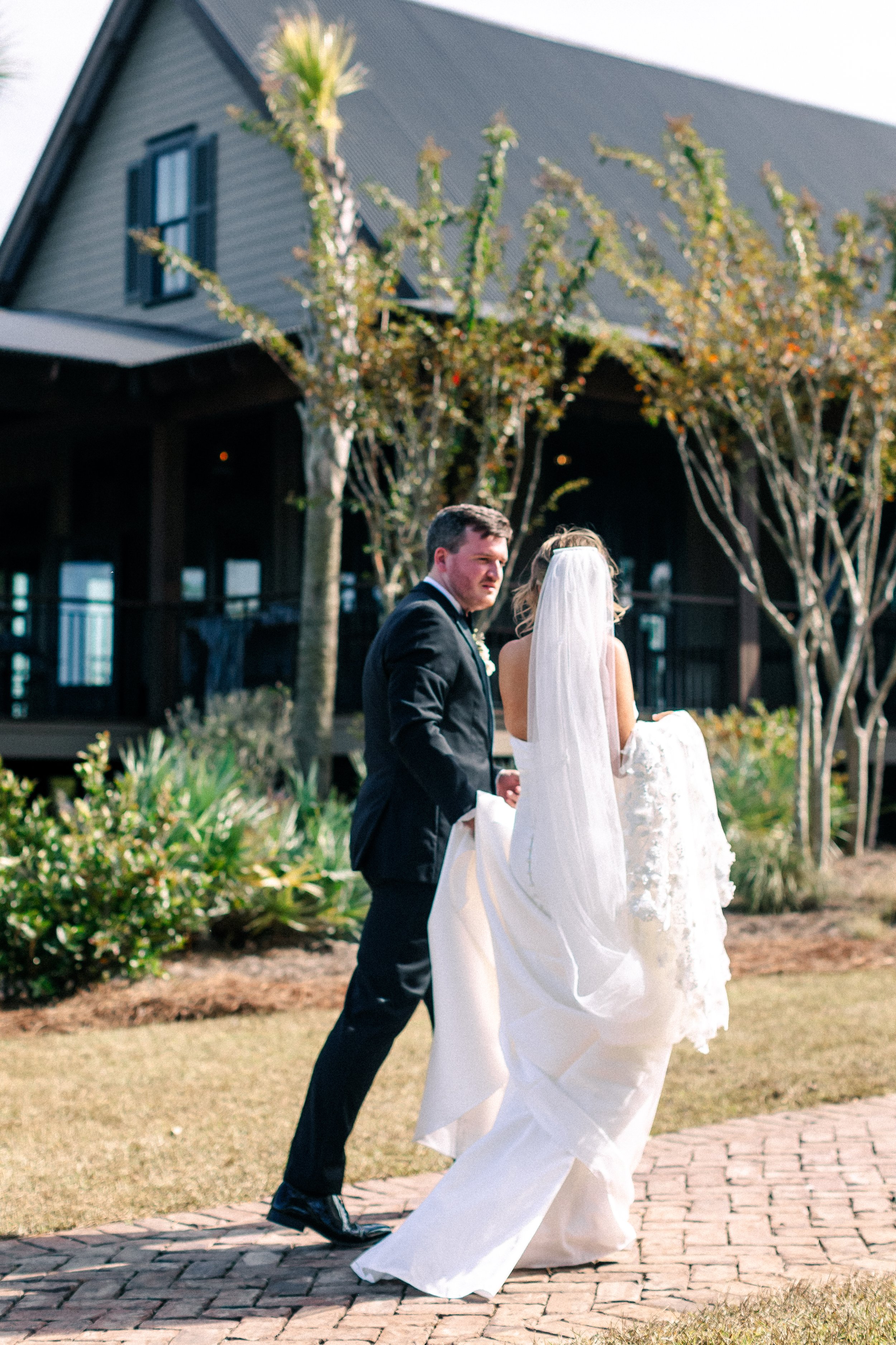 A bride and groom walking outdoors, with the groom in a black suit and the bride in a white wedding dress and veil, near a house with trees.