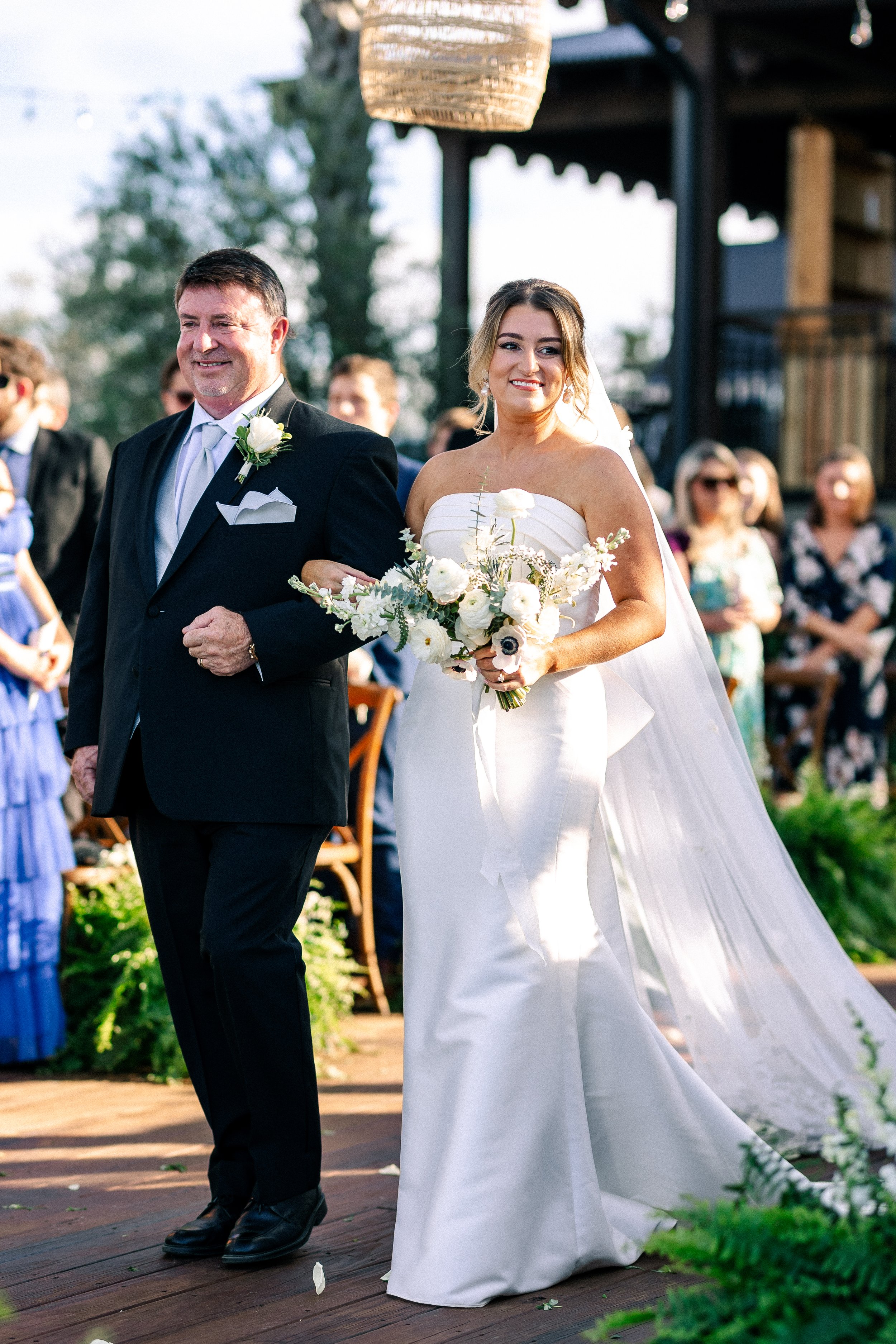 A bride walking down the aisle arm-in-arm with a man, possibly her father, during an outdoor wedding ceremony. The bride is holding a bouquet of white flowers and wearing a strapless white wedding gown with a long veil. The man is dressed in a black 