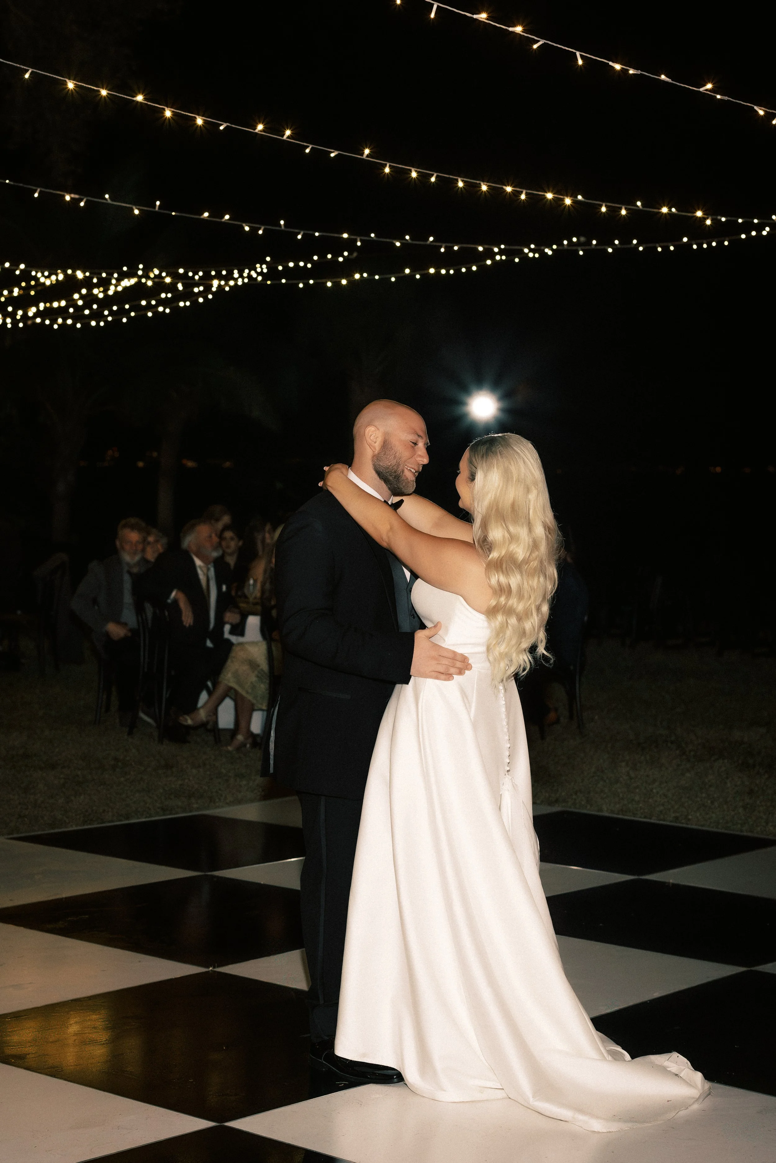 A bride and groom share a first dance at night under string lights and a full moon, with guests seated in the background.